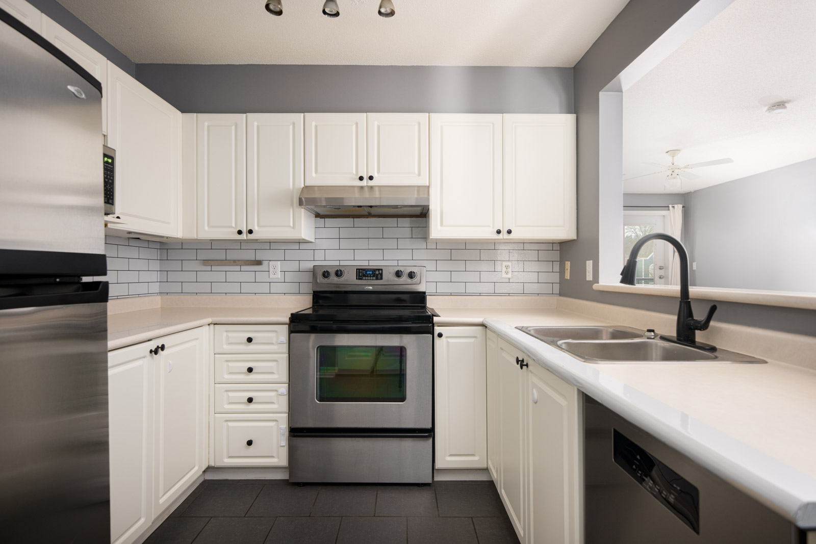 Modern kitchen with white cabinets, stainless steel appliances, black tile floor, subway tile backsplash, and a double sink under a large open pass-through window.