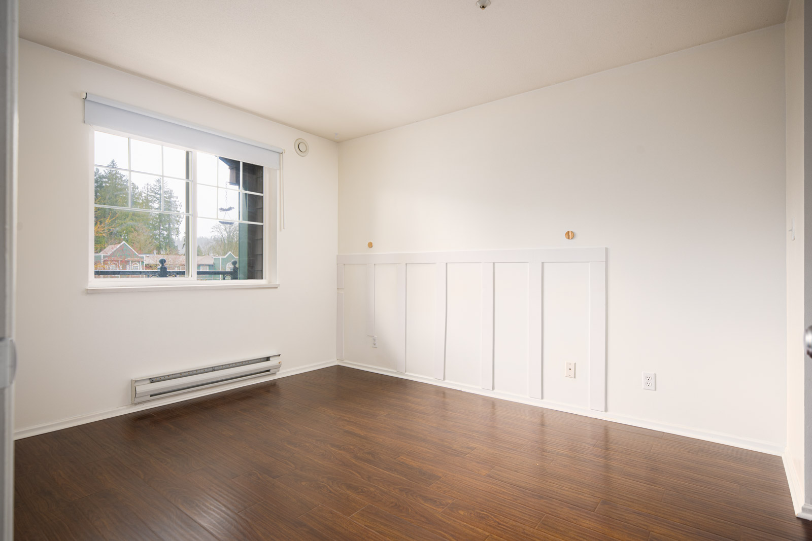 Empty room with white walls, large window, white trim, baseboard heater, and dark wood flooring. Daylight streams in through the window.