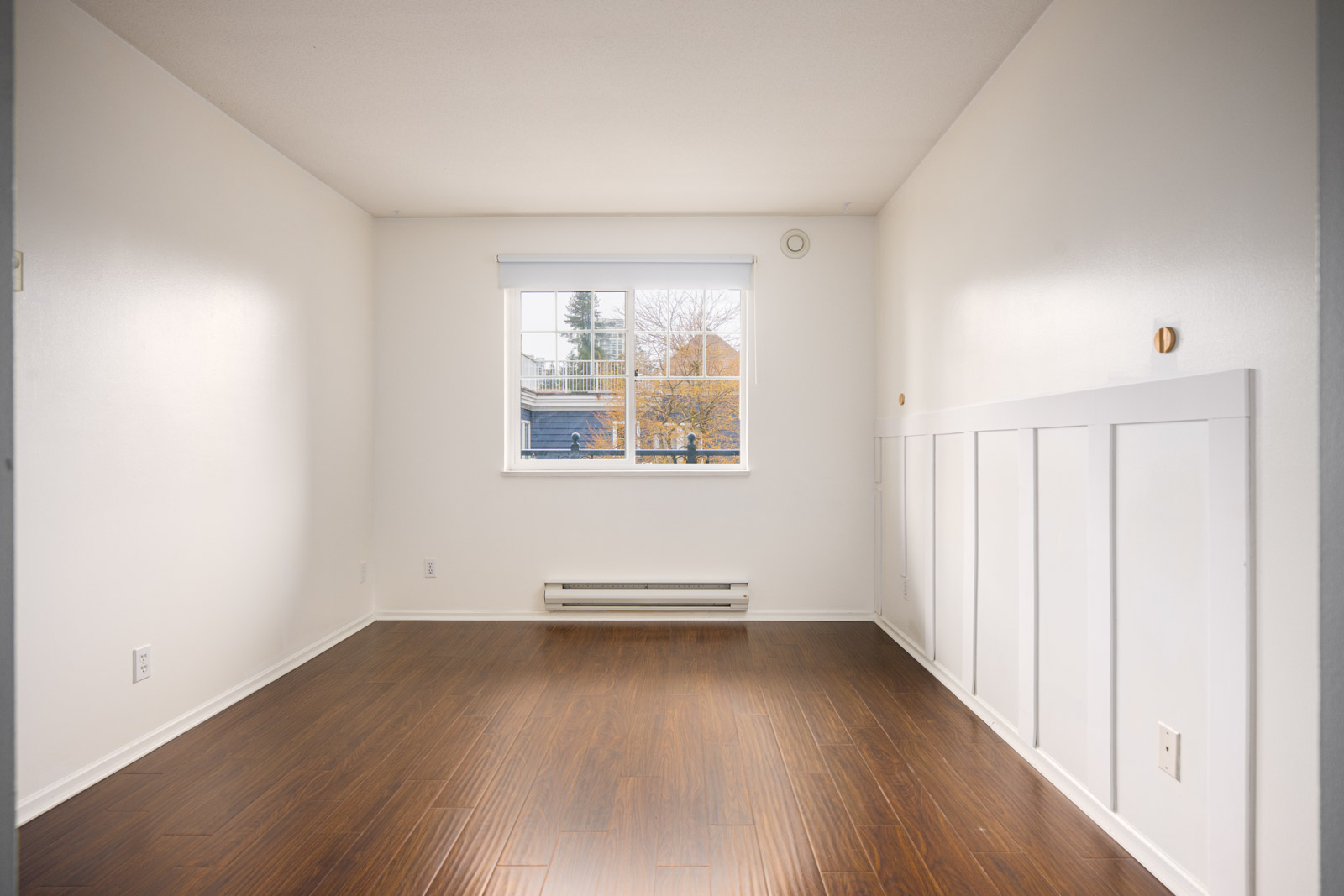 Empty room with white walls, wooden floor, a single window with a view of trees and rooftops, and a baseboard heater below the window.