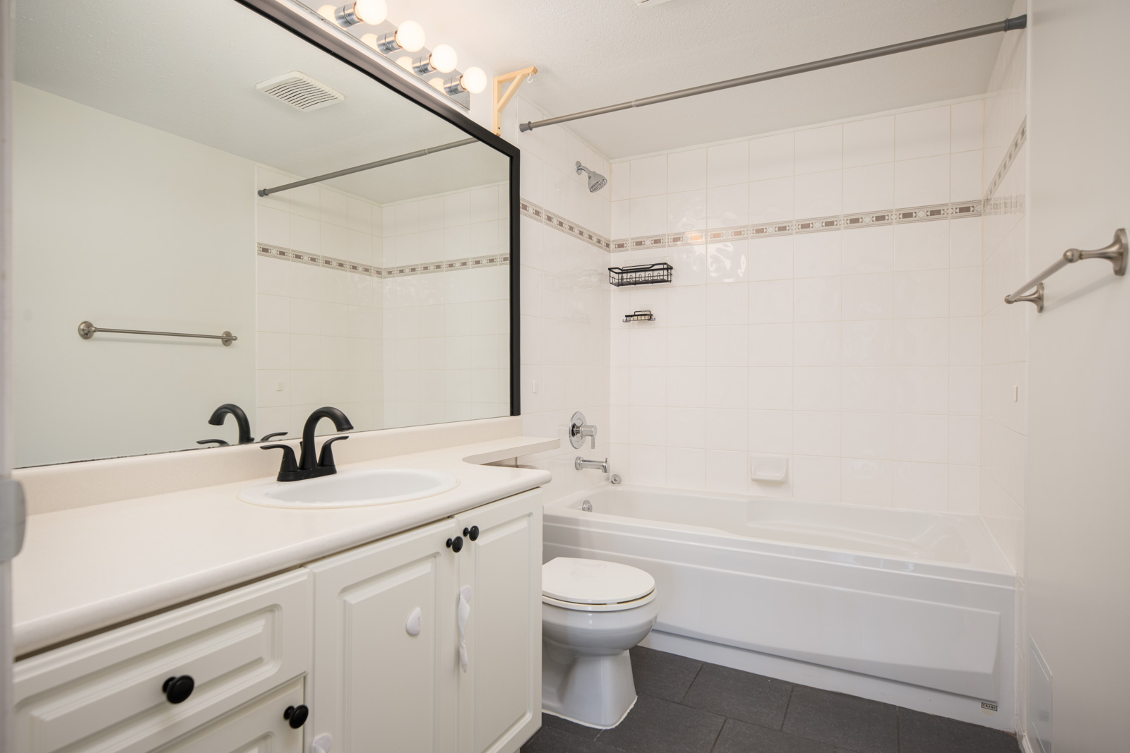 A clean, white bathroom with a vanity, black fixtures, a toilet, and a bathtub with a tiled shower surround under bright lights.