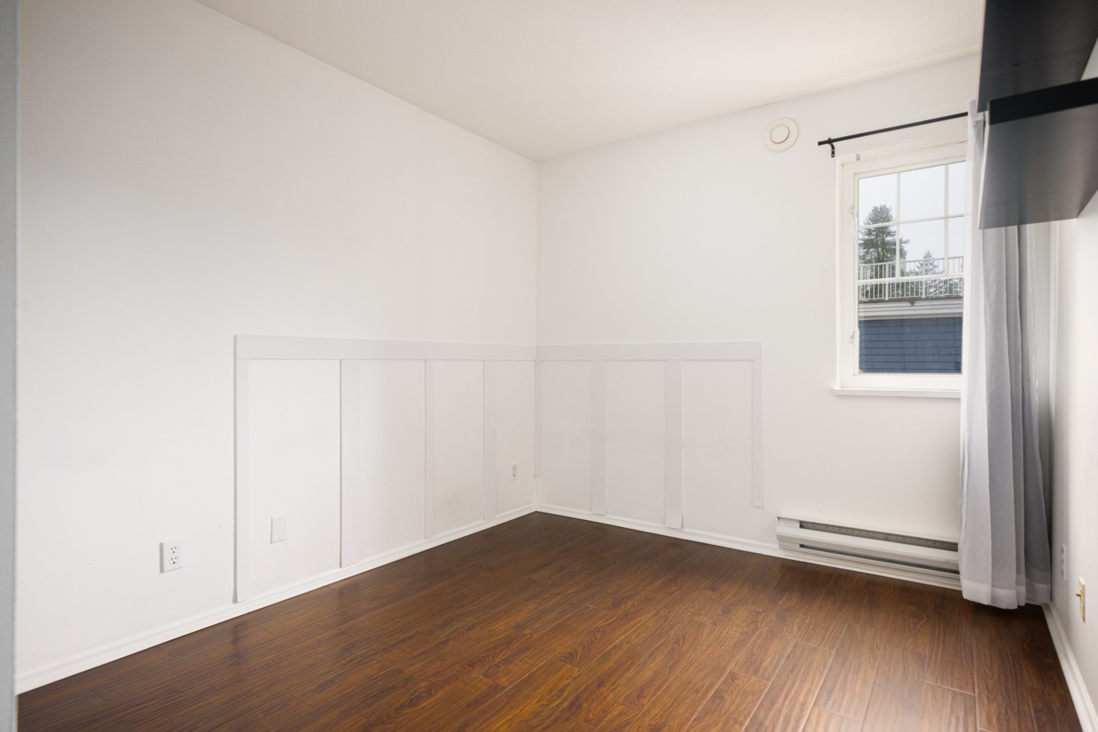 Empty room with white walls, dark wood flooring, a window with sheer curtain, baseboard heater, and a wall-mounted black shelf. Natural light enters from the window.