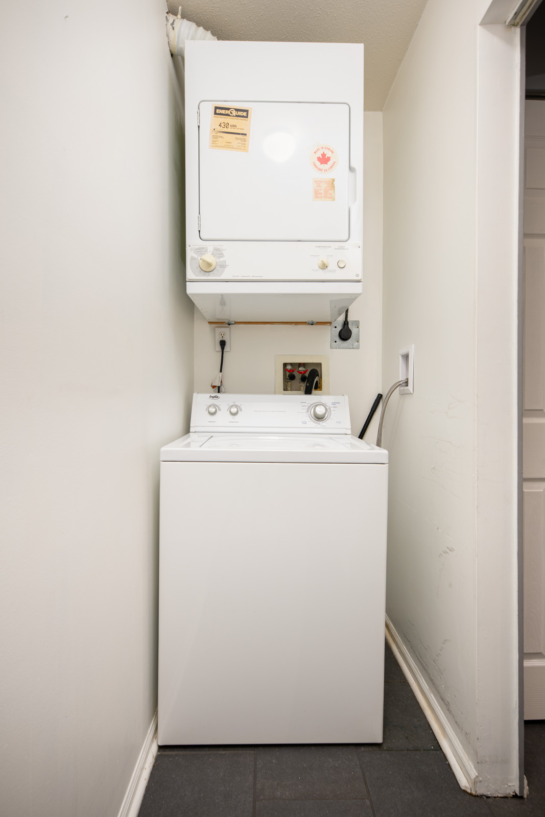 A stacked white washer and dryer set is placed in a small, narrow laundry area with tile flooring and off-white walls.