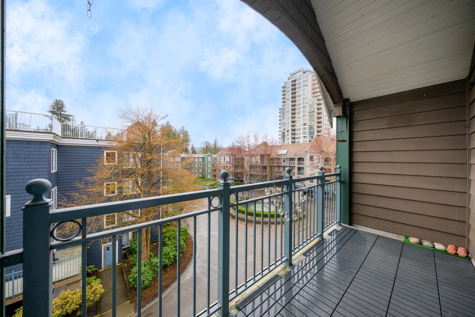 View from a covered apartment balcony with railing, overlooking a courtyard, trees, and several multi-story residential buildings under a partly cloudy sky.