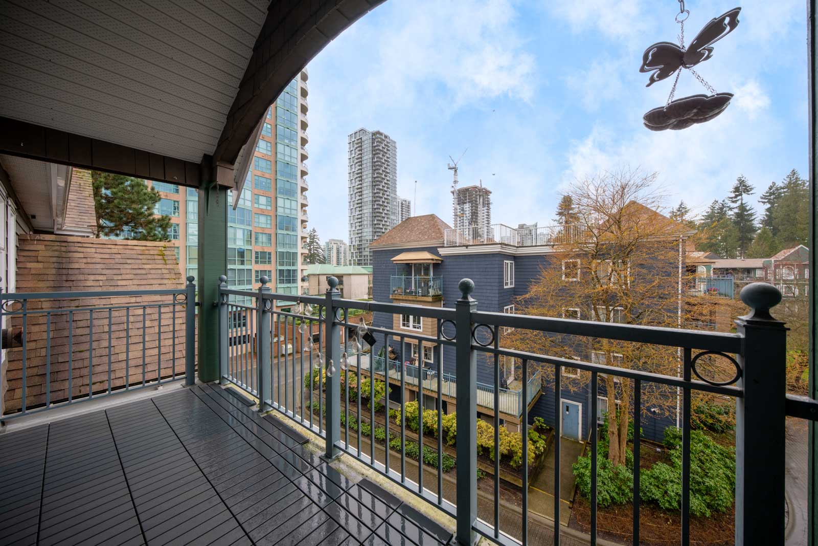 A covered balcony with metal railings overlooks a residential area and high-rise buildings, with trees and cloudy sky in the background.