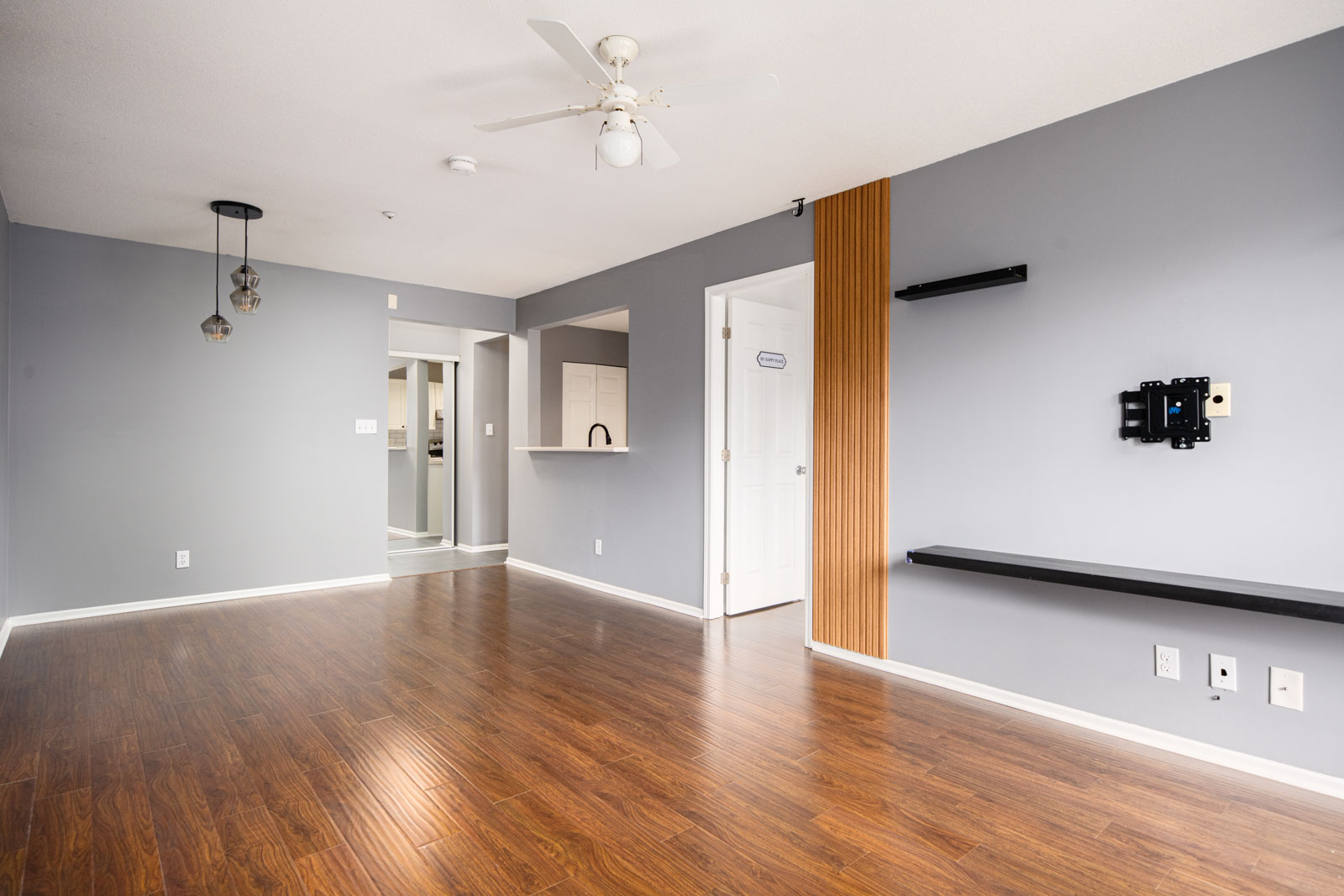 Empty living room with wood flooring, gray walls, ceiling fan, and open pass-through window to kitchen; wall shelves and TV mount are installed on one wall.