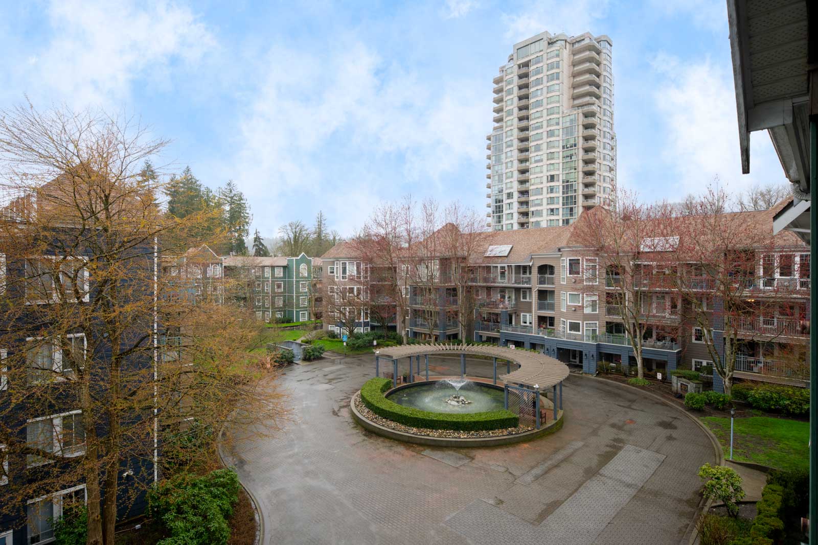 A residential apartment complex with a central courtyard featuring a round fountain, surrounded by multi-story buildings and a tall high-rise in the background.