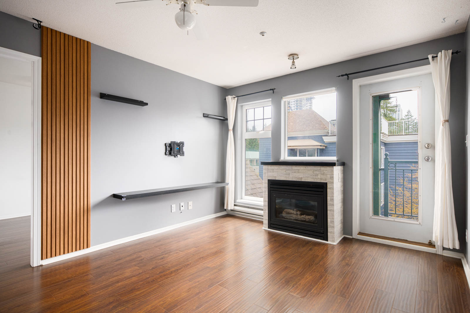A modern living room with light gray walls, wood flooring, a wall-mounted TV bracket, floating shelves, a fireplace, two windows, and a glass door with white curtains.
