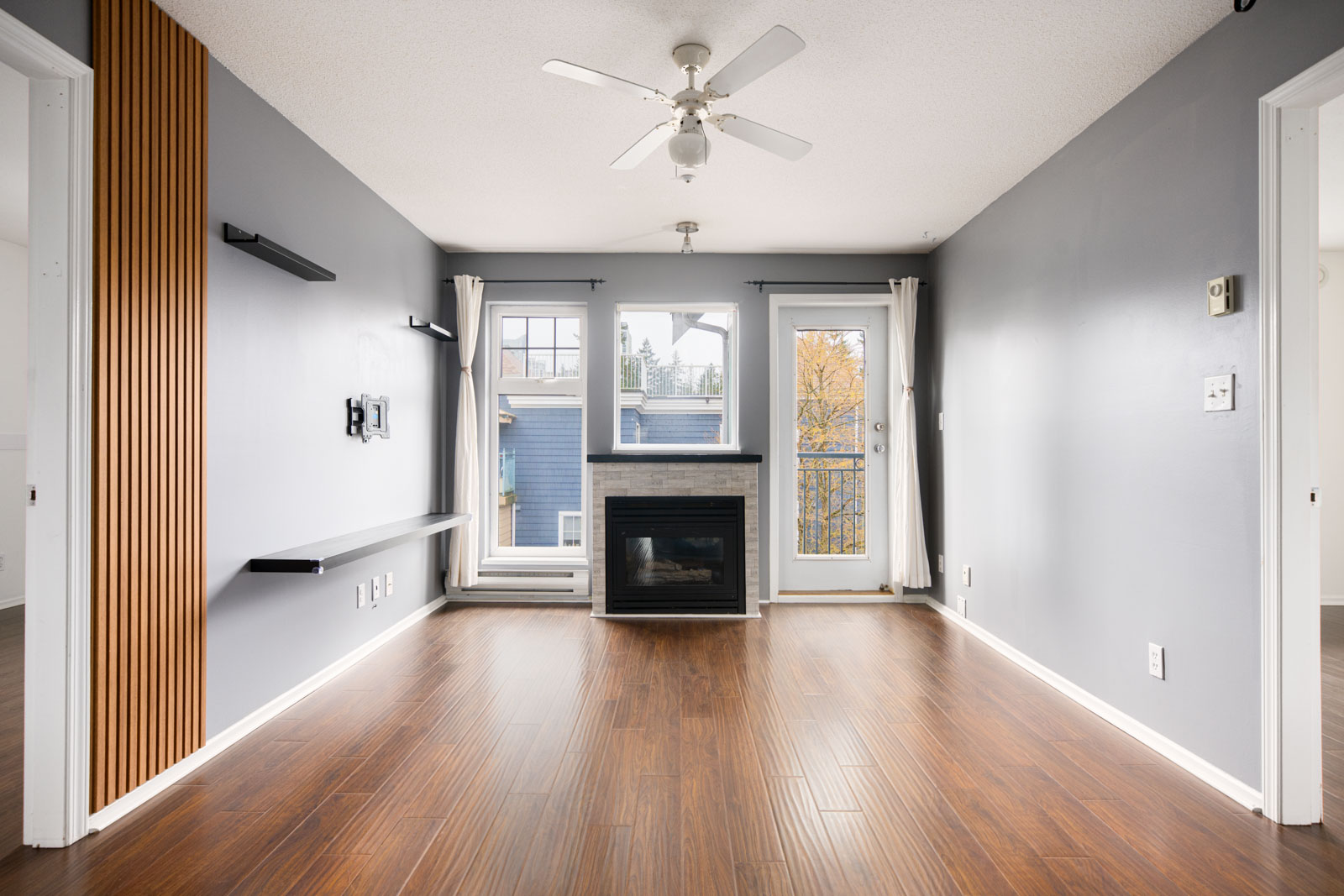 Empty living room with gray walls, wood floors, a ceiling fan, fireplace, wall shelves, and door to a small balcony with a view of nearby buildings and trees.