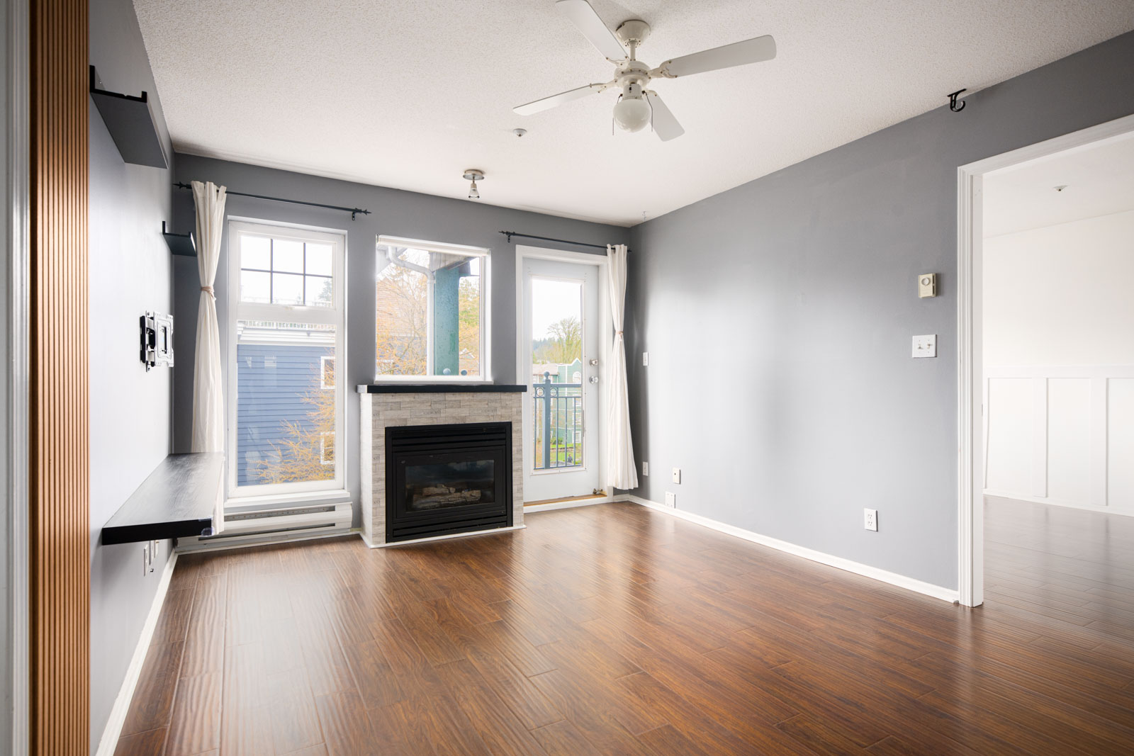 Empty living room with gray walls, wood flooring, a ceiling fan, wall-mounted shelf, fireplace, and glass doors leading to a small balcony; a bedroom is visible through an open doorway.