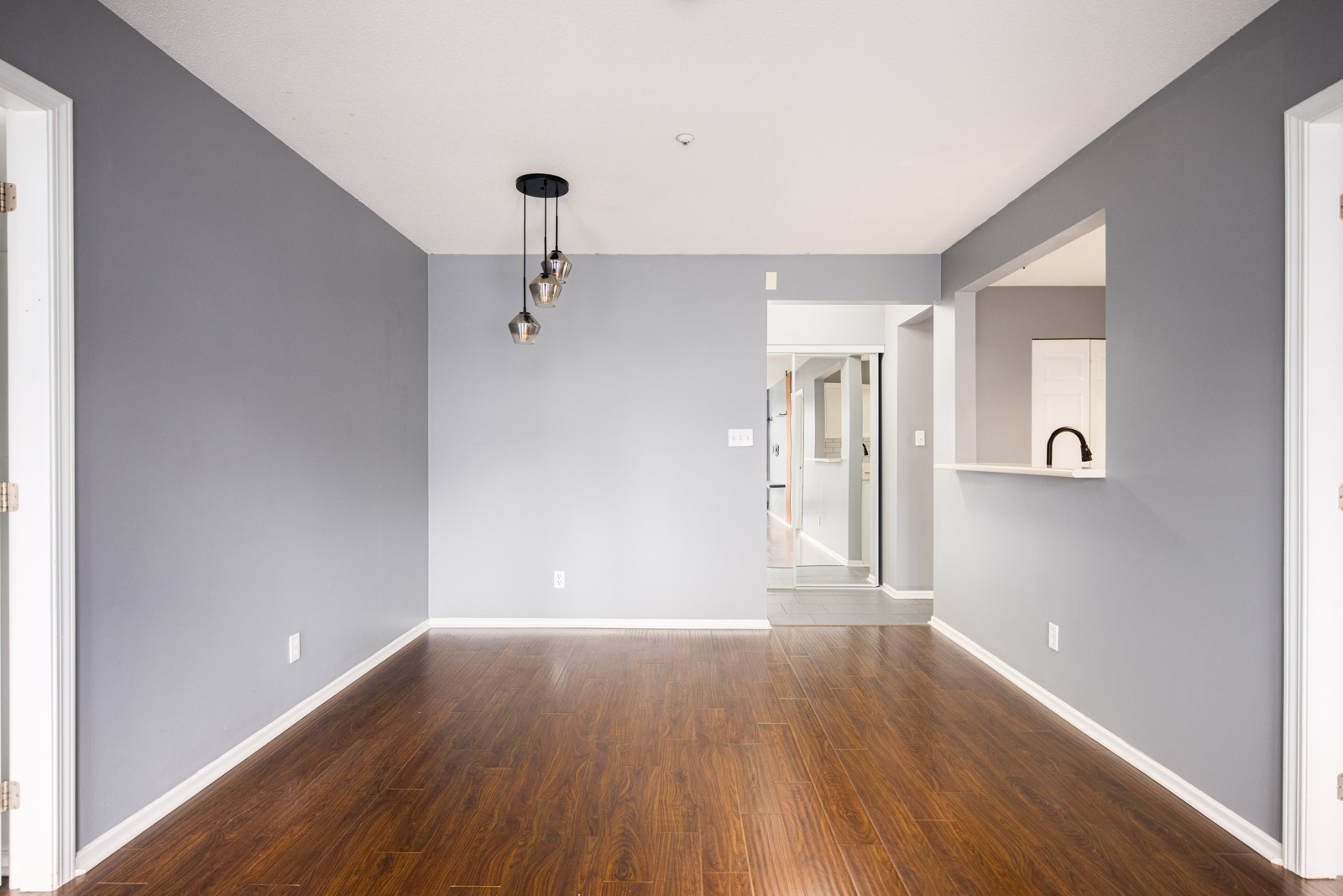 Empty room with gray walls, pendant lights, and dark wood flooring, featuring a pass-through window to the kitchen and an open doorway leading to a hallway.