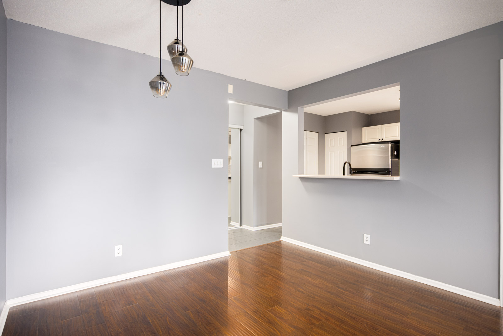 Empty room with gray walls, dark wood floor, three pendant lights, and an open window to a kitchen with stainless steel appliances.