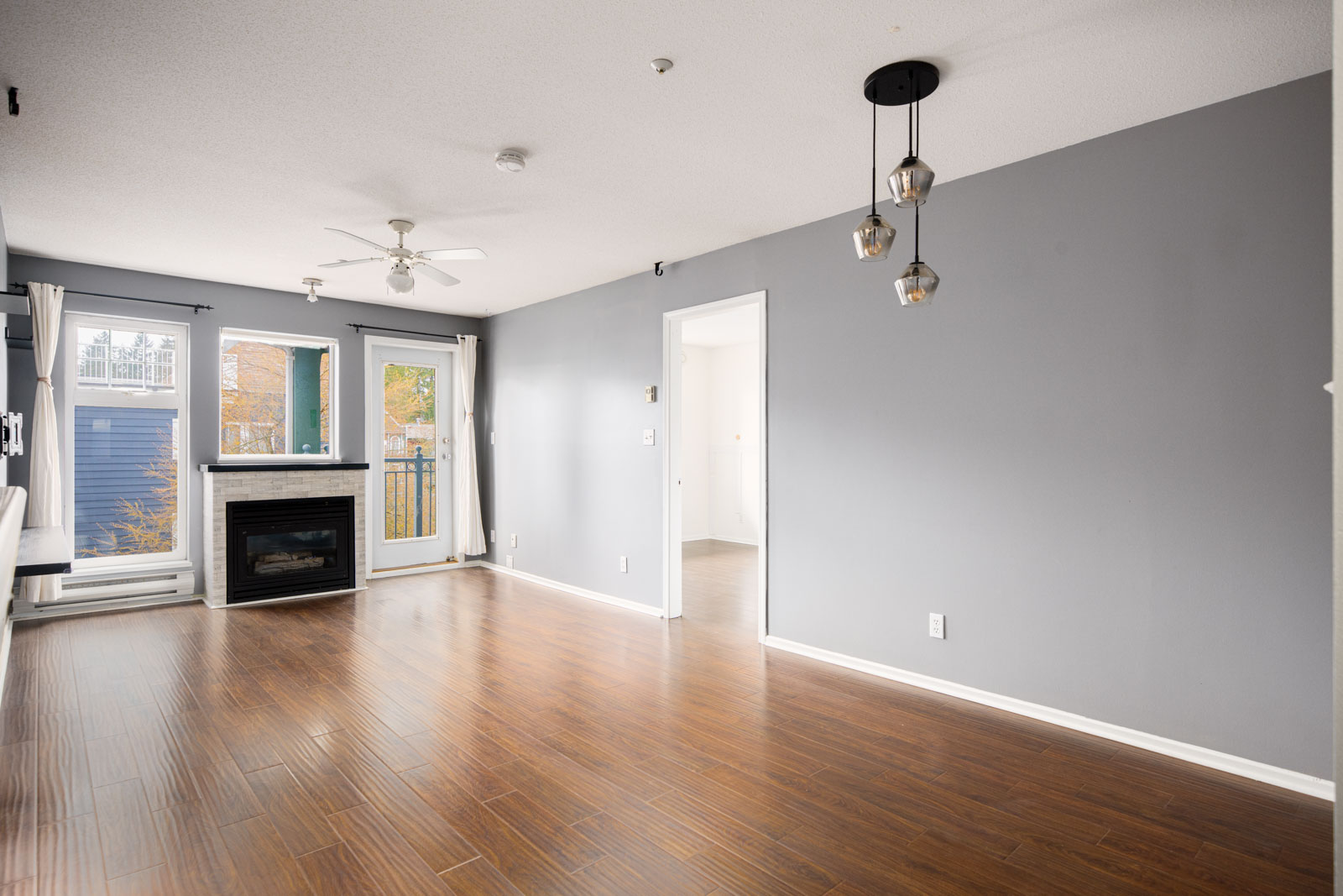 A modern, unfurnished living room with gray walls, wood flooring, a ceiling fan, corner fireplace, large windows, and hanging light fixtures. Doorway leads to another room.