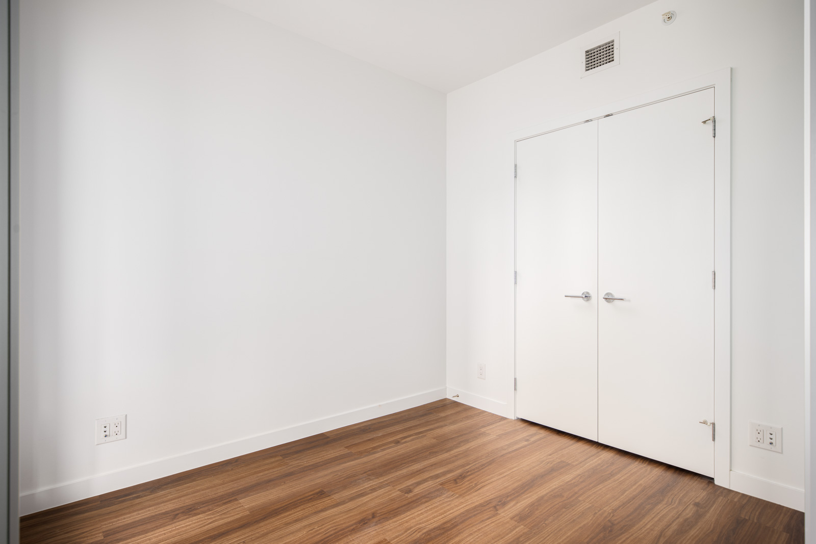 Empty room with white walls, wooden floor, and closed double white doors. An air vent and electrical outlets are visible on the walls.