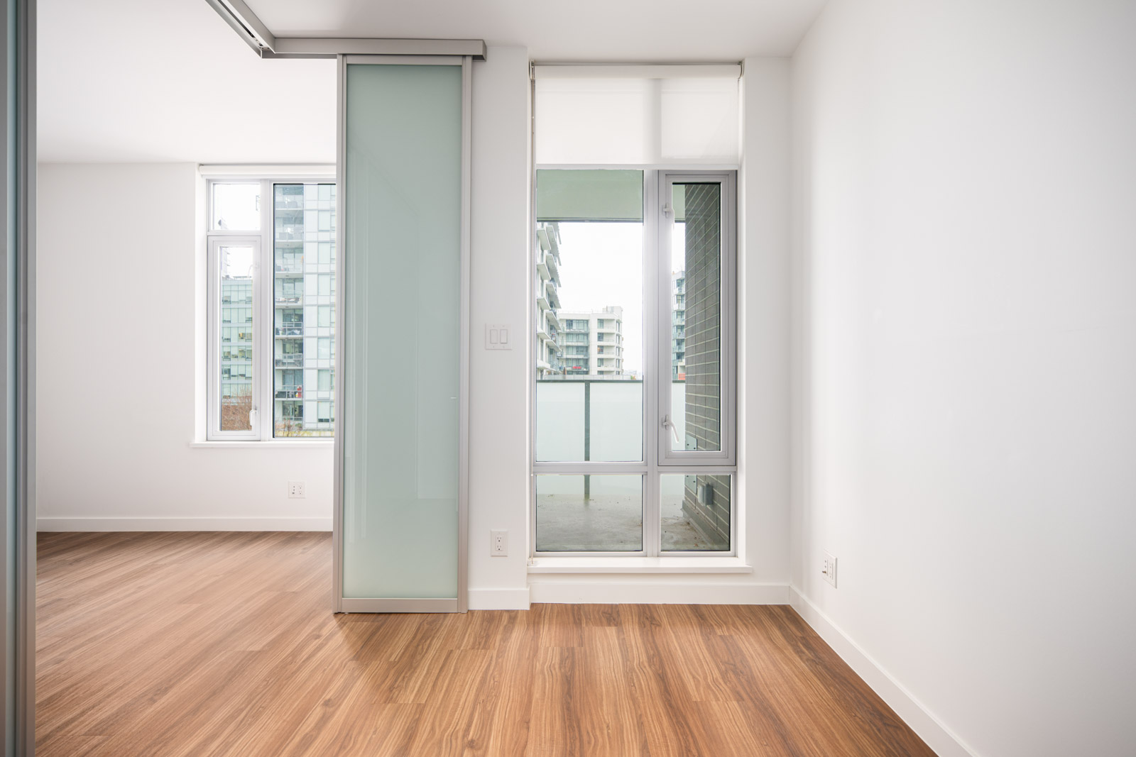 A bright, empty room with wood flooring, large windows, a glass sliding door, and a view of apartment buildings outside.