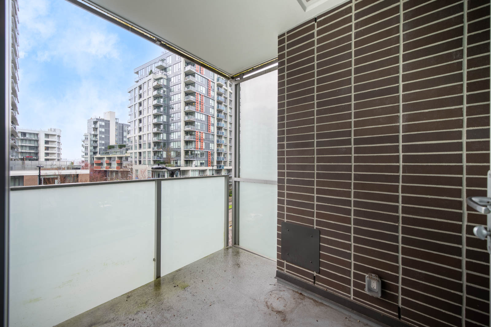 Modern apartment balcony with frosted glass railing, brown brick wall, and view of surrounding residential high-rise buildings under a partly cloudy sky.