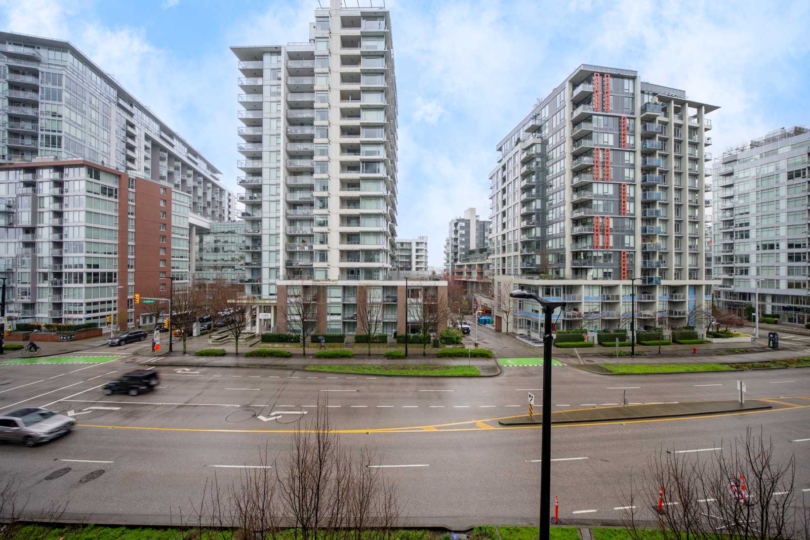 Wide street with moving vehicles and bicycles, bordered by modern high-rise apartment buildings under a partly cloudy sky.