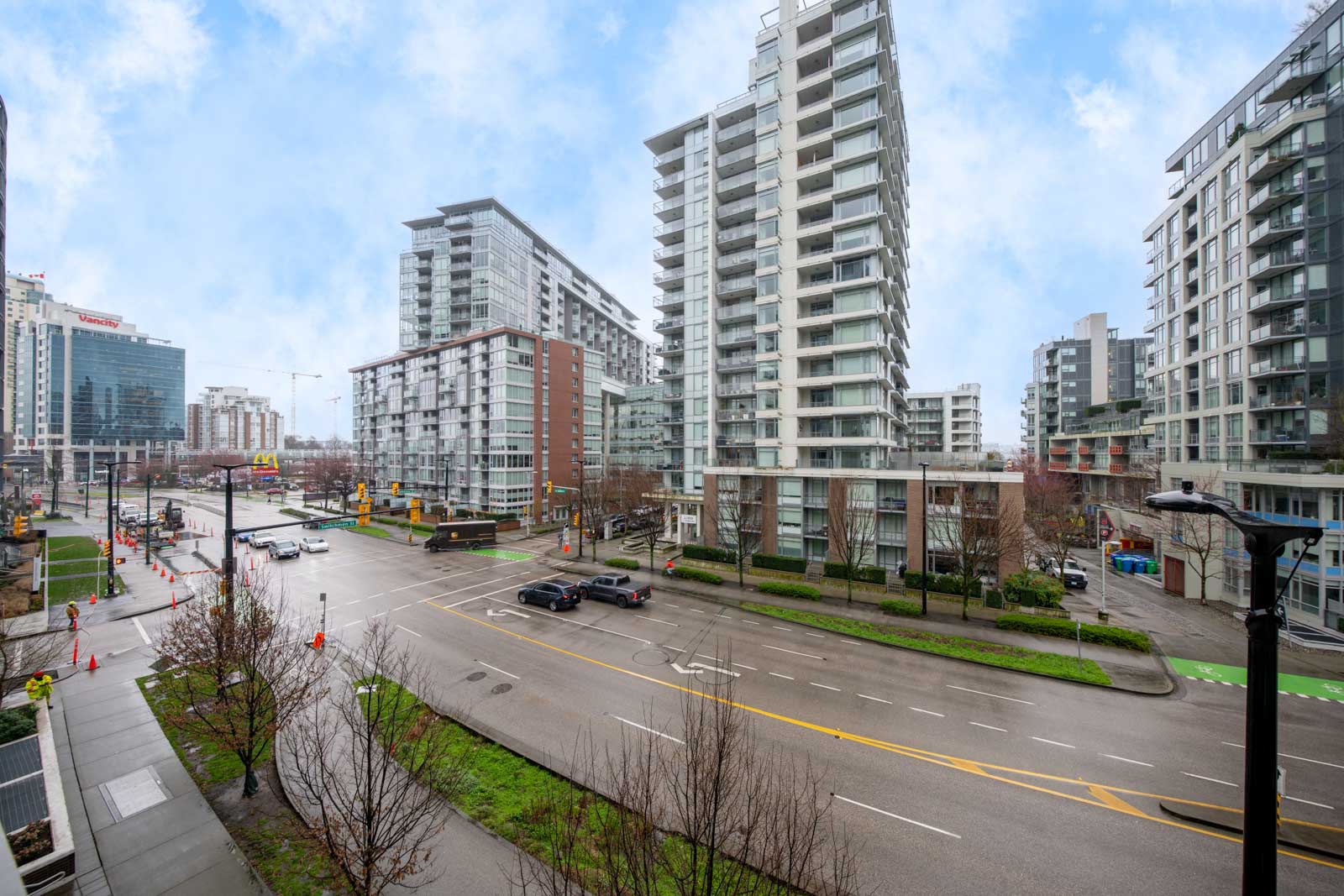 View of a city intersection with modern high-rise buildings, several cars on the road, and overcast skies. The sidewalks are lined with bare trees and some greenery.