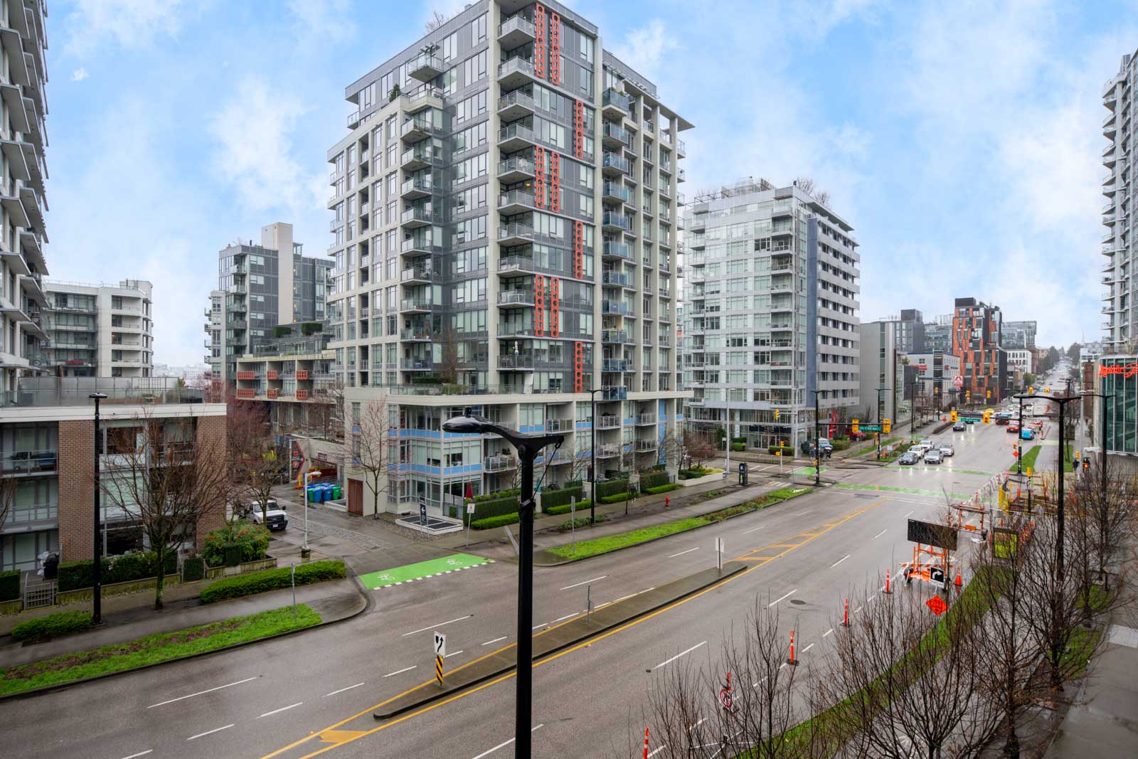 Street view of a modern city with mid-rise apartment buildings, empty sidewalks, light traffic, and cloudy sky.