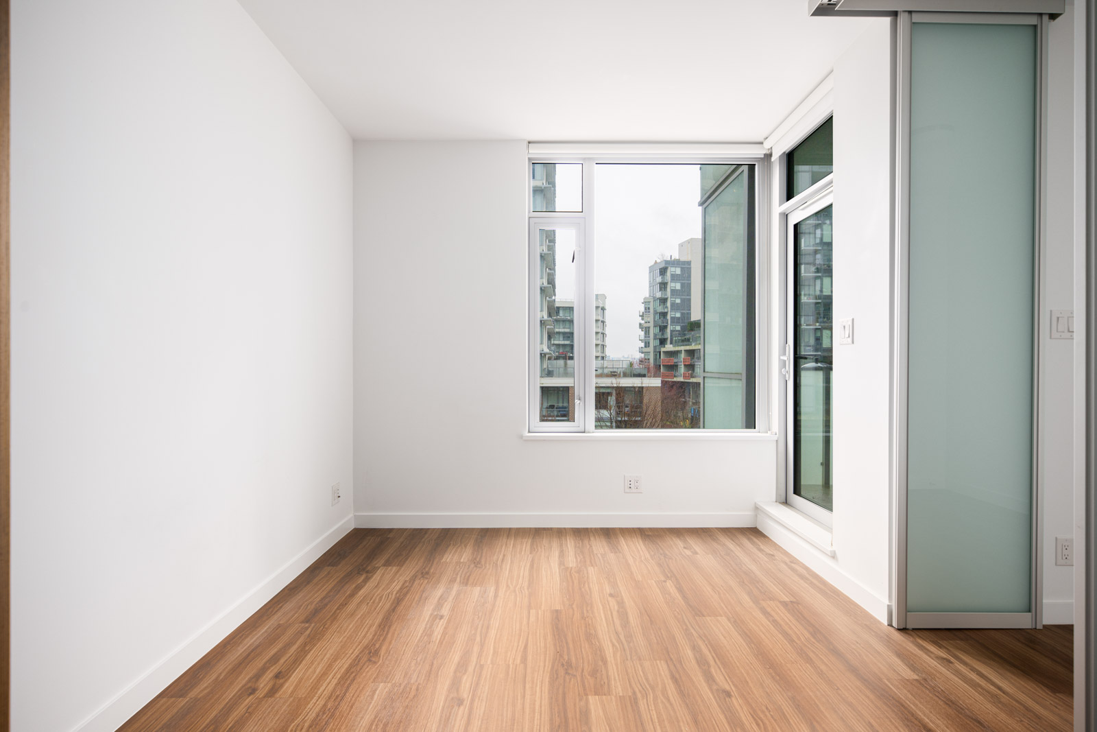 Empty modern room with white walls, wood flooring, a large window showing city buildings, and a glass sliding door on the right.