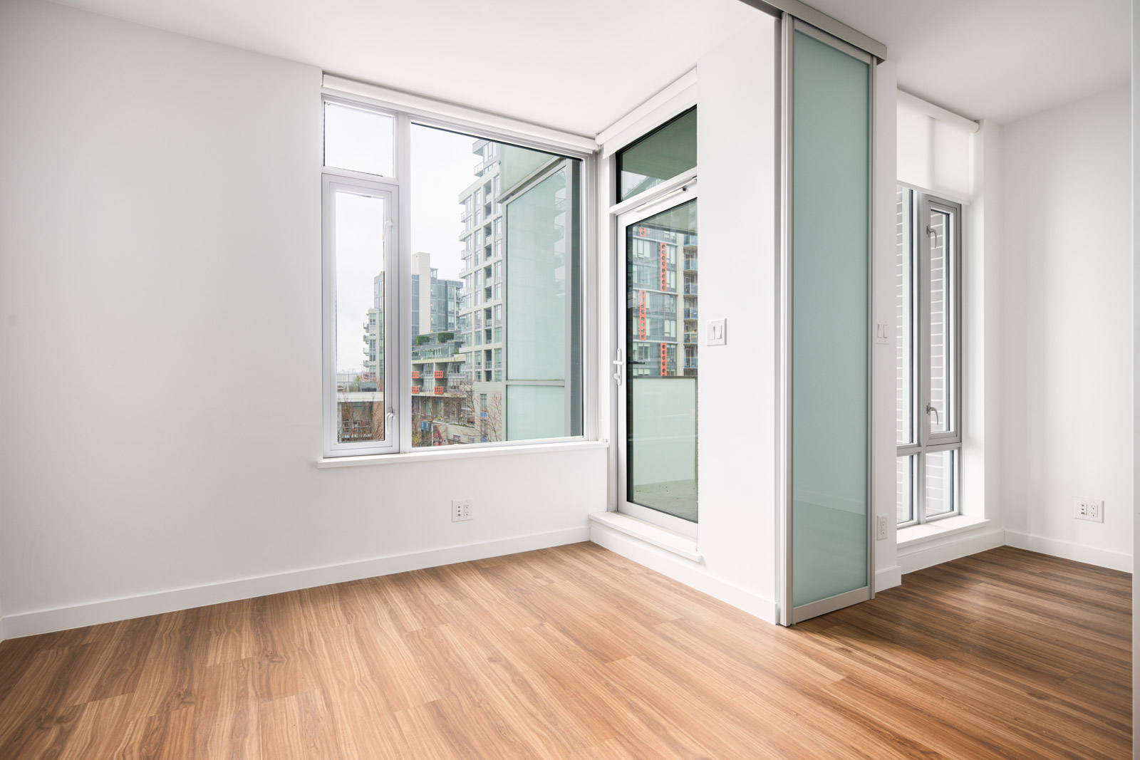 Unfurnished modern apartment room with wood flooring, large windows, sliding glass door to a balcony, and white walls. Urban buildings are visible outside.