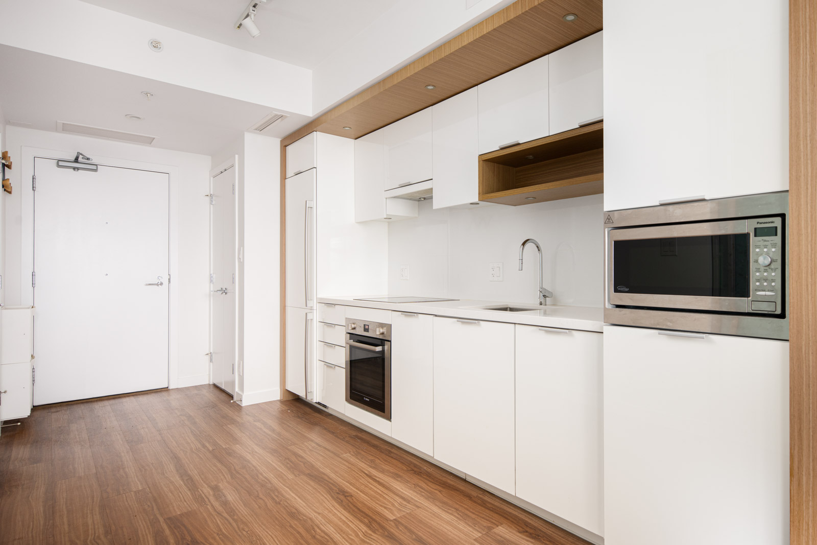 Modern kitchen with white cabinets, built-in oven, microwave, and sink; wood flooring and minimal decor in a compact, open space near an entrance door.