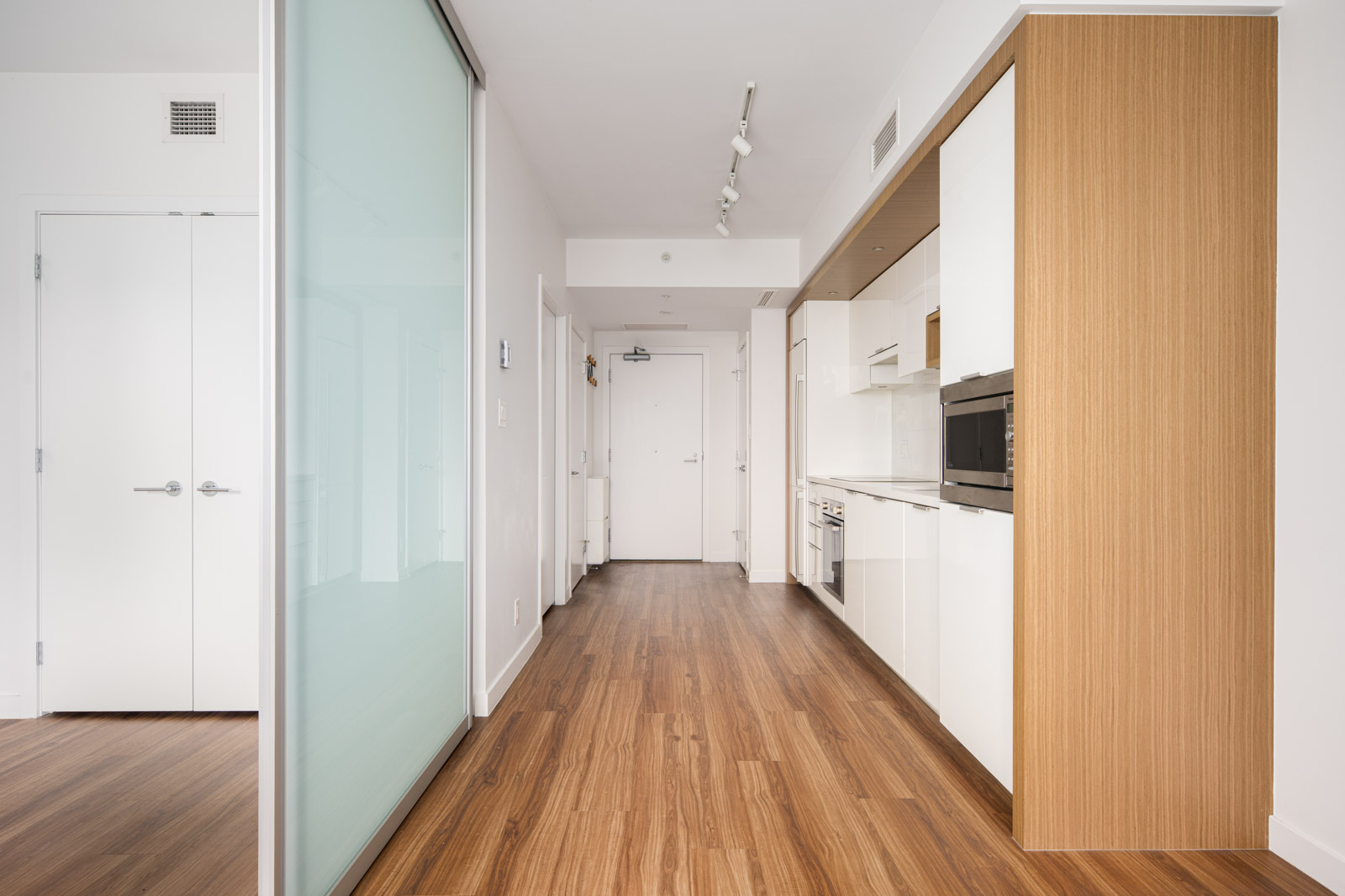 A modern, narrow kitchen with white cabinets, wood flooring, built-in appliances, and sliding glass doors leading to a separate room.