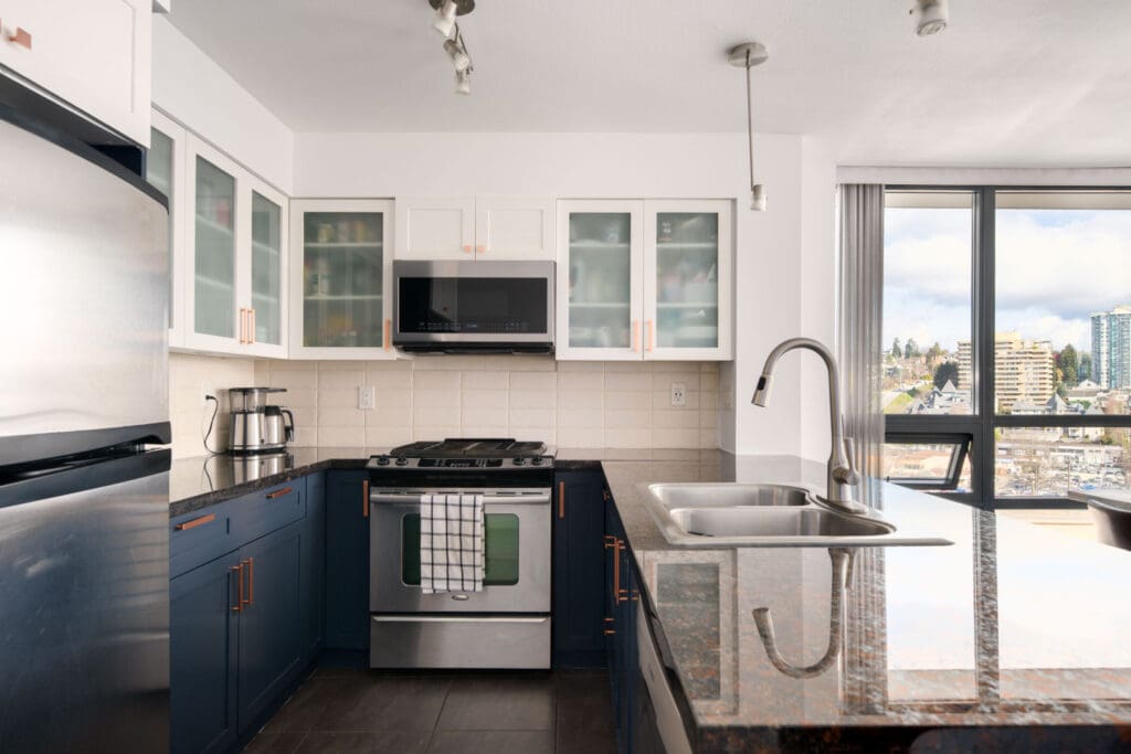 Modern kitchen with stainless steel appliances, blue lower cabinets, white upper cabinets with glass doors, a double sink, and large windows overlooking a cityscape.