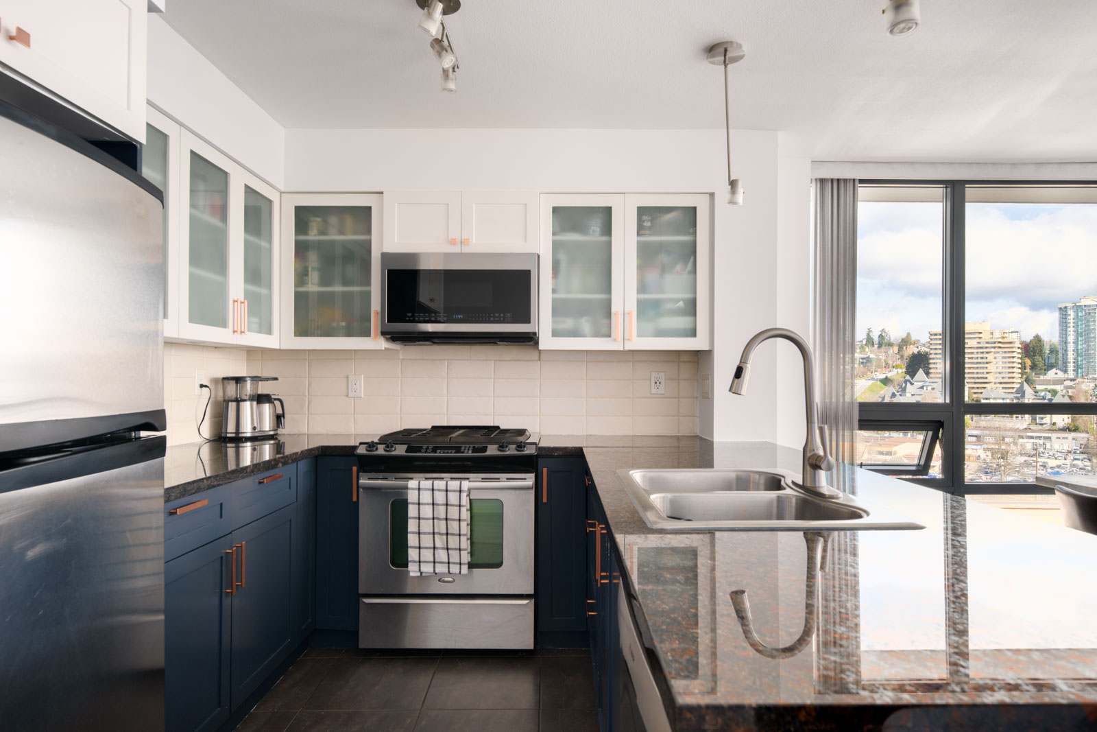 Modern kitchen with stainless steel appliances, blue lower cabinets, white upper cabinets with glass doors, a double sink, and large windows overlooking a cityscape.