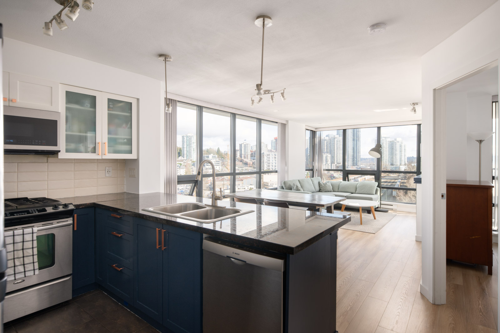 Modern open-plan kitchen and living area with large windows, city skyline views, dark countertops, and light wood flooring.