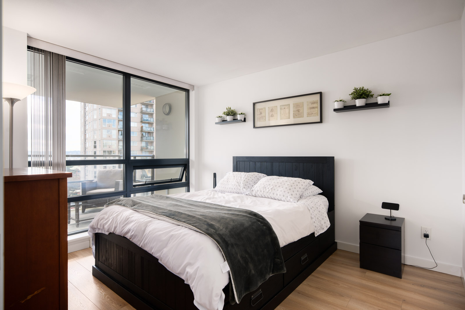 A modern bedroom with a large window, a black bed with storage drawers, white bedding, two floating shelves with plants, and a nightstand.
