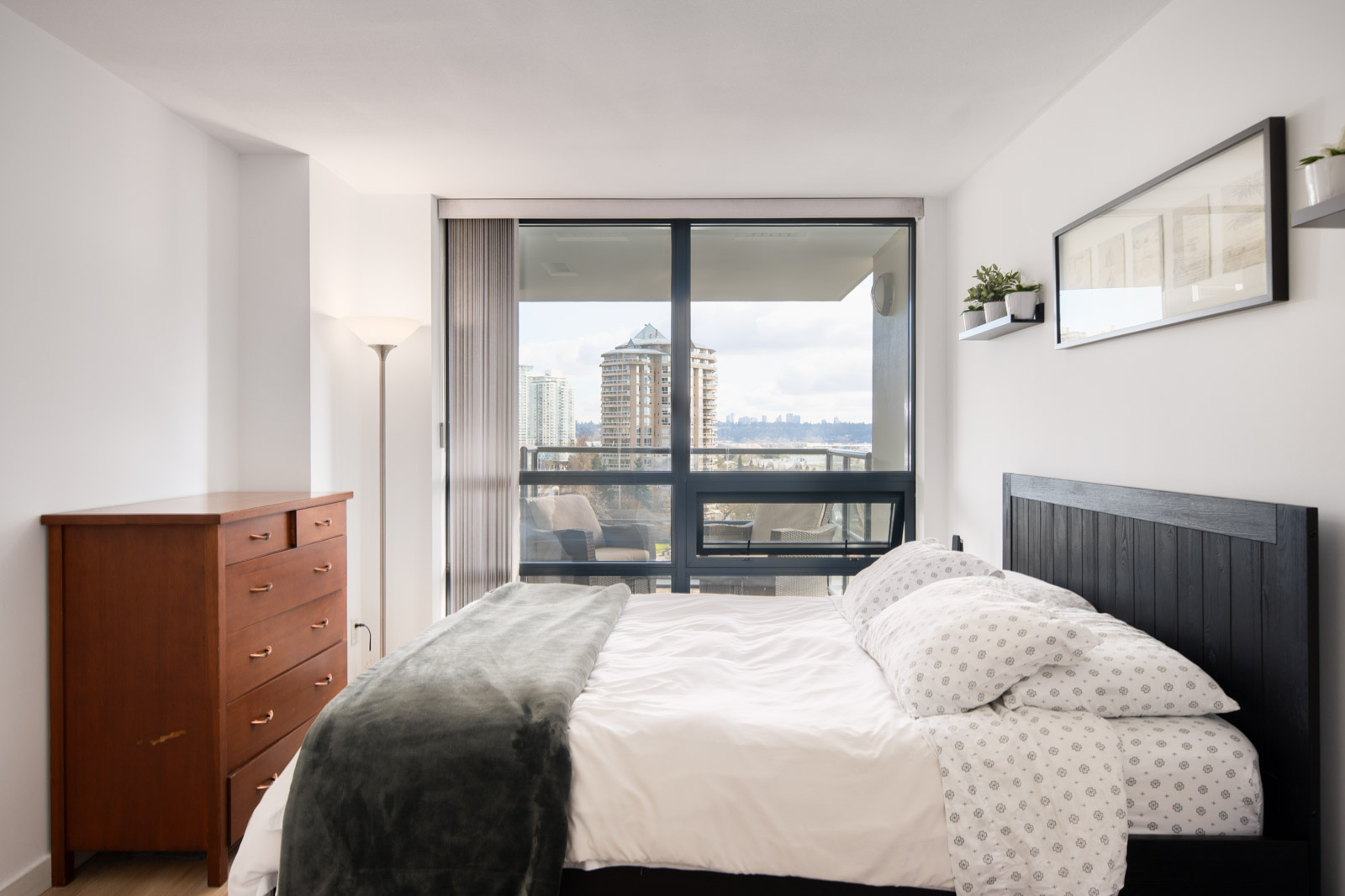 Modern bedroom with a black bed frame, white bedding, wooden dresser, and large window showing a city view and balcony.