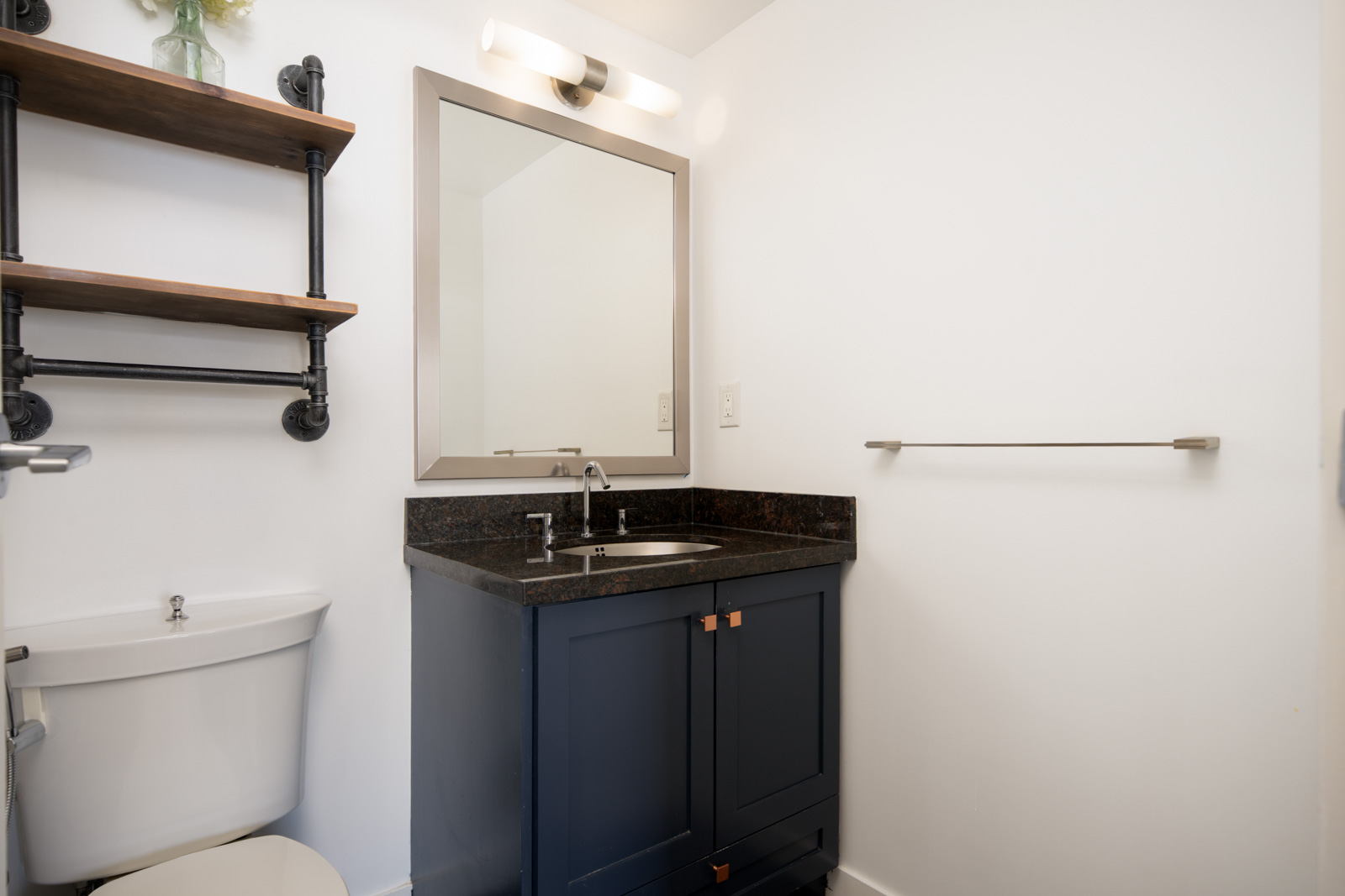 A small bathroom with a navy blue vanity, black countertop, wall-mounted mirror, toilet, open shelves, and a towel bar on a white wall.