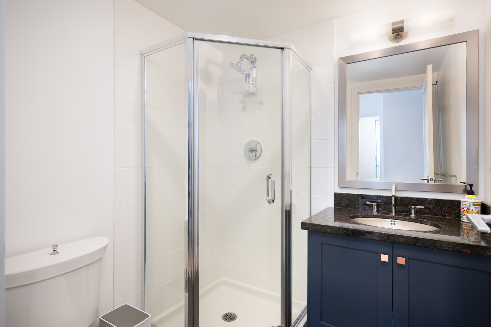 Modern bathroom with a corner glass shower, a white toilet, a sink with a dark countertop and blue cabinet, and a wall mirror above the sink.