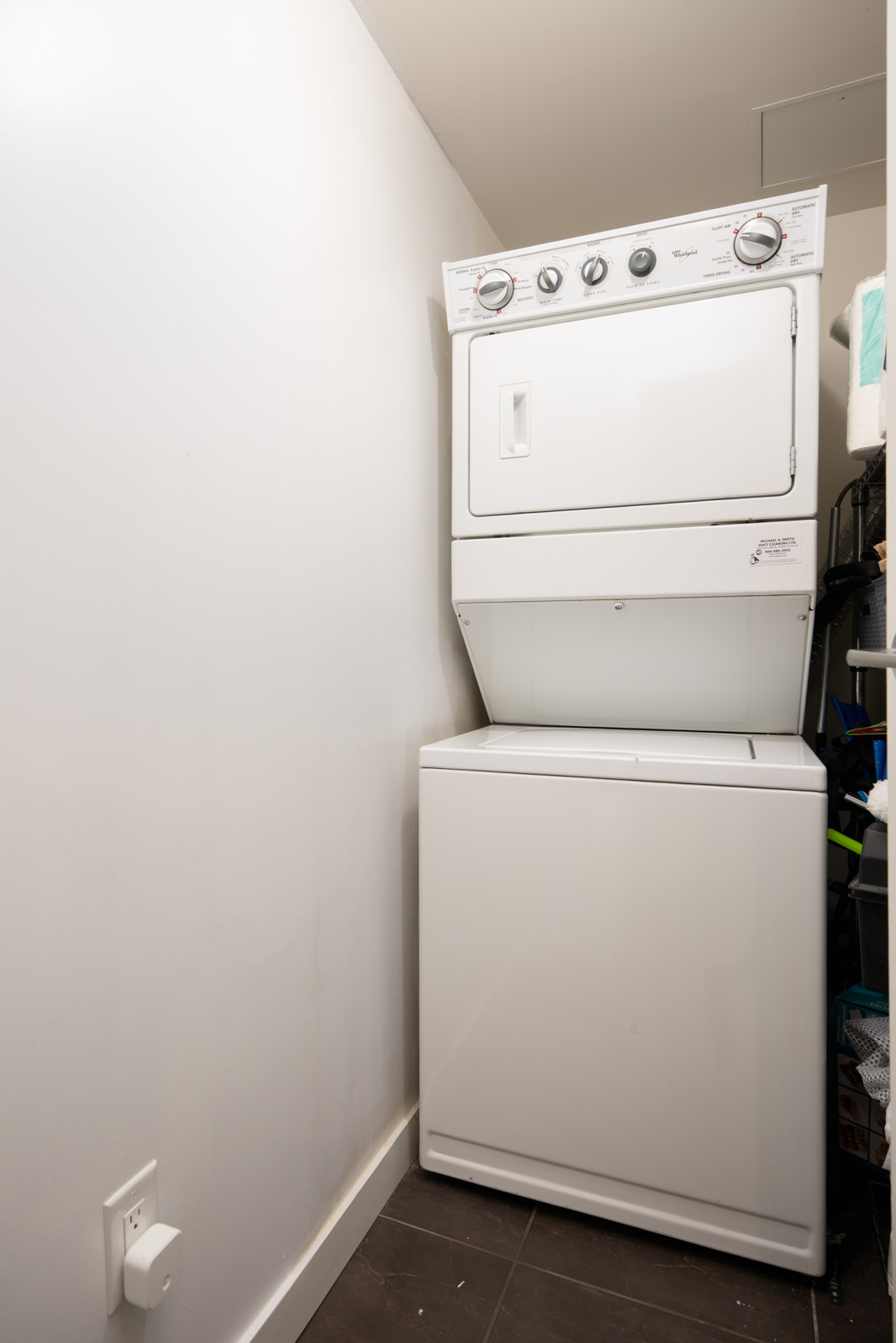 A white stacked washer and dryer unit positioned in a narrow laundry room with white walls and dark flooring.