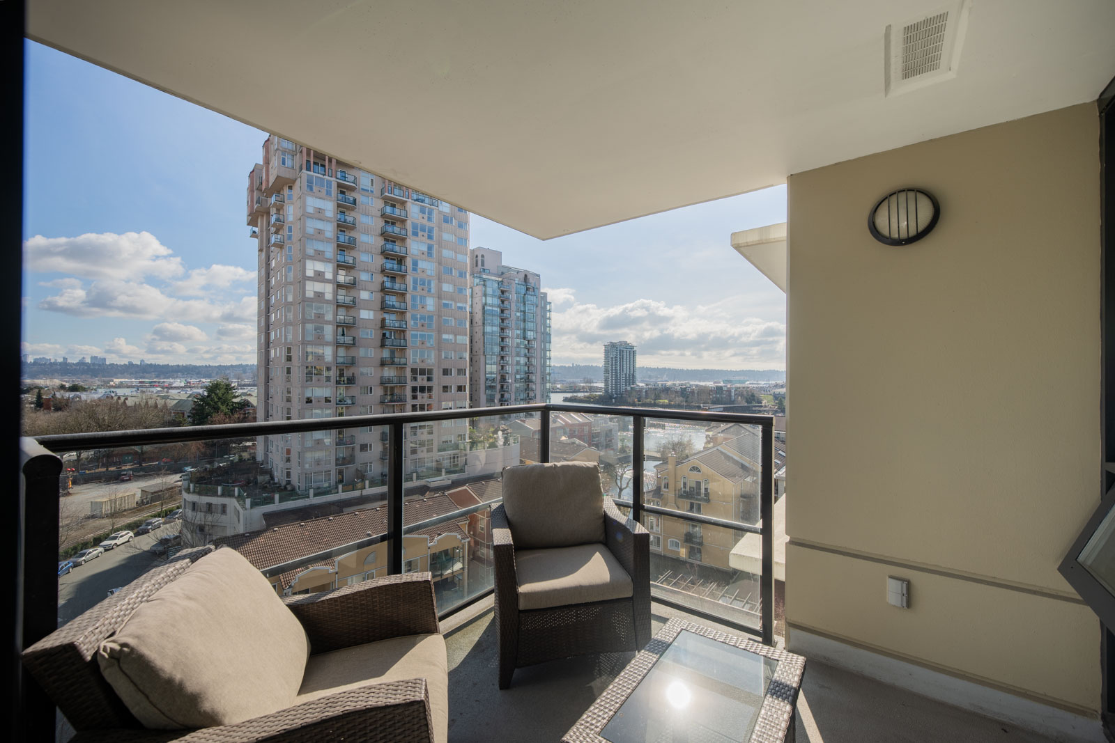 A furnished balcony with two chairs and a glass-top table overlooks city buildings and a distant landscape under a partly cloudy sky.