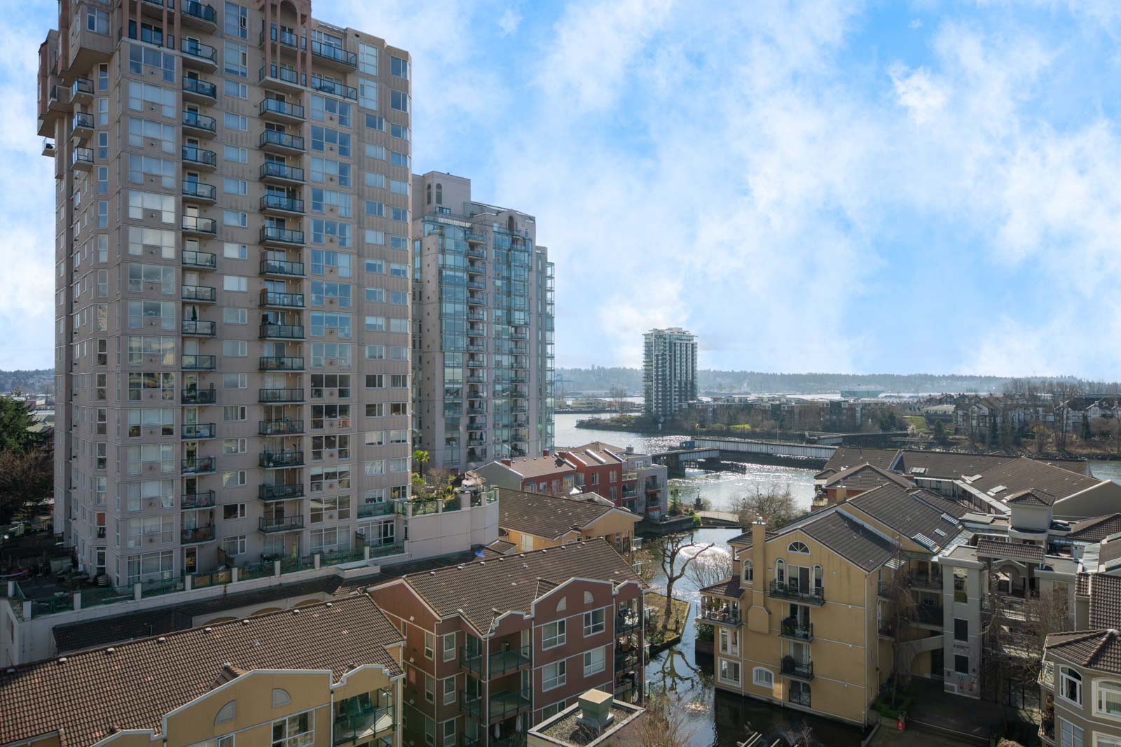 High-rise apartment buildings and colorful townhouses are shown alongside a canal, with a clear sky and distant waterway visible in the background.