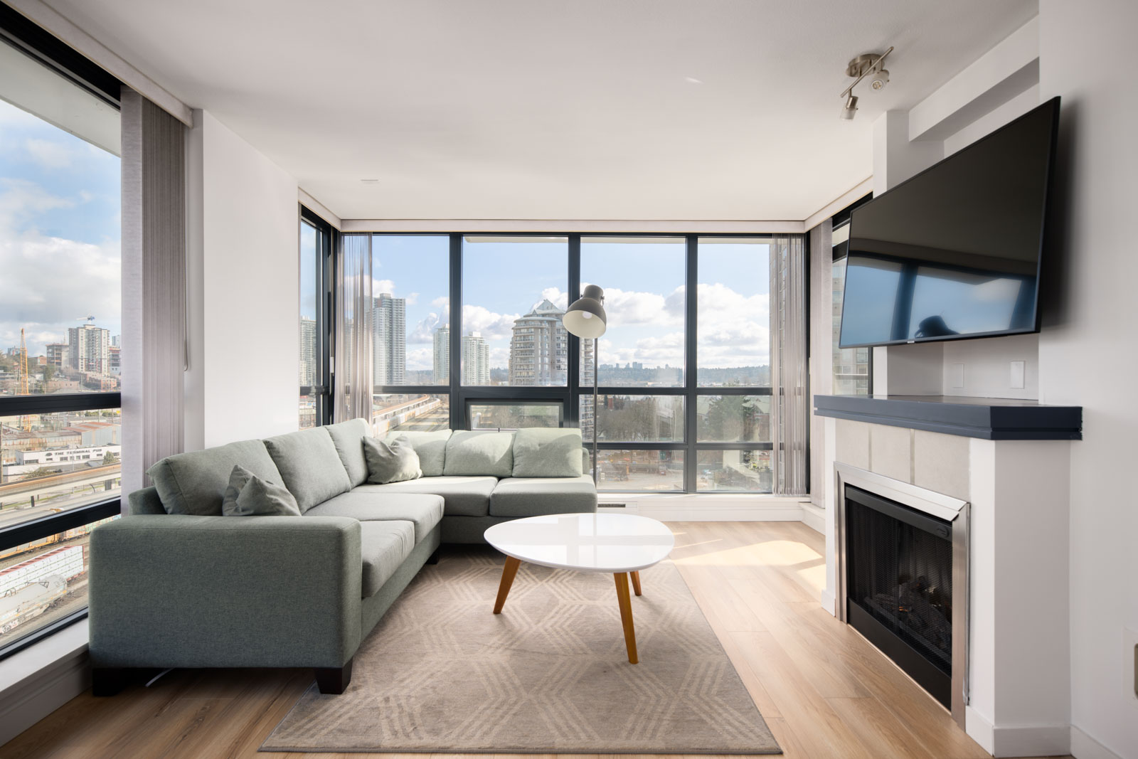 Modern living room with large windows, light green sectional sofa, round white coffee table, fireplace, wall-mounted TV, and city view outside.