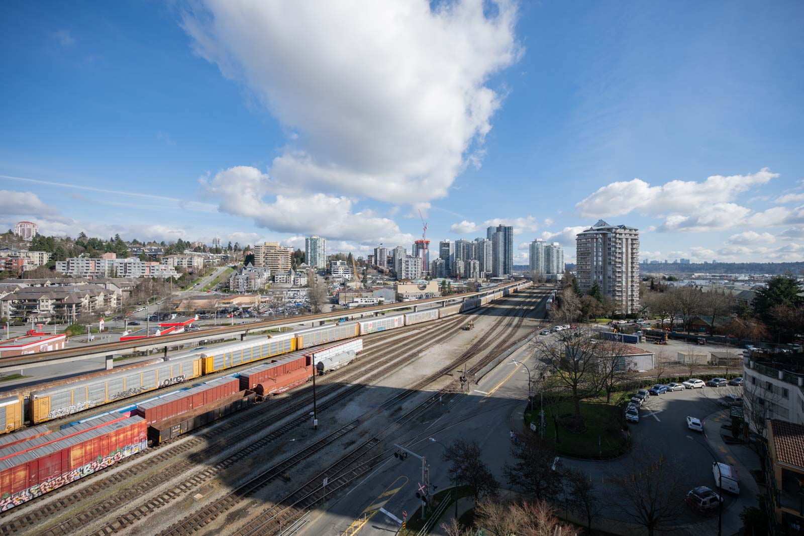 A cityscape with train tracks, freight trains, and buildings under a blue sky with scattered clouds. Trees and parking areas border the tracks.