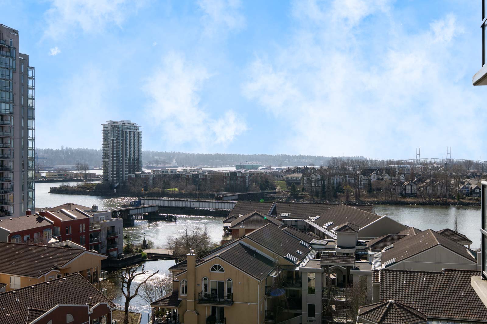 View of a river with residential buildings in the foreground, a bridge, and mid-rise towers on the opposite bank under a partly cloudy sky.