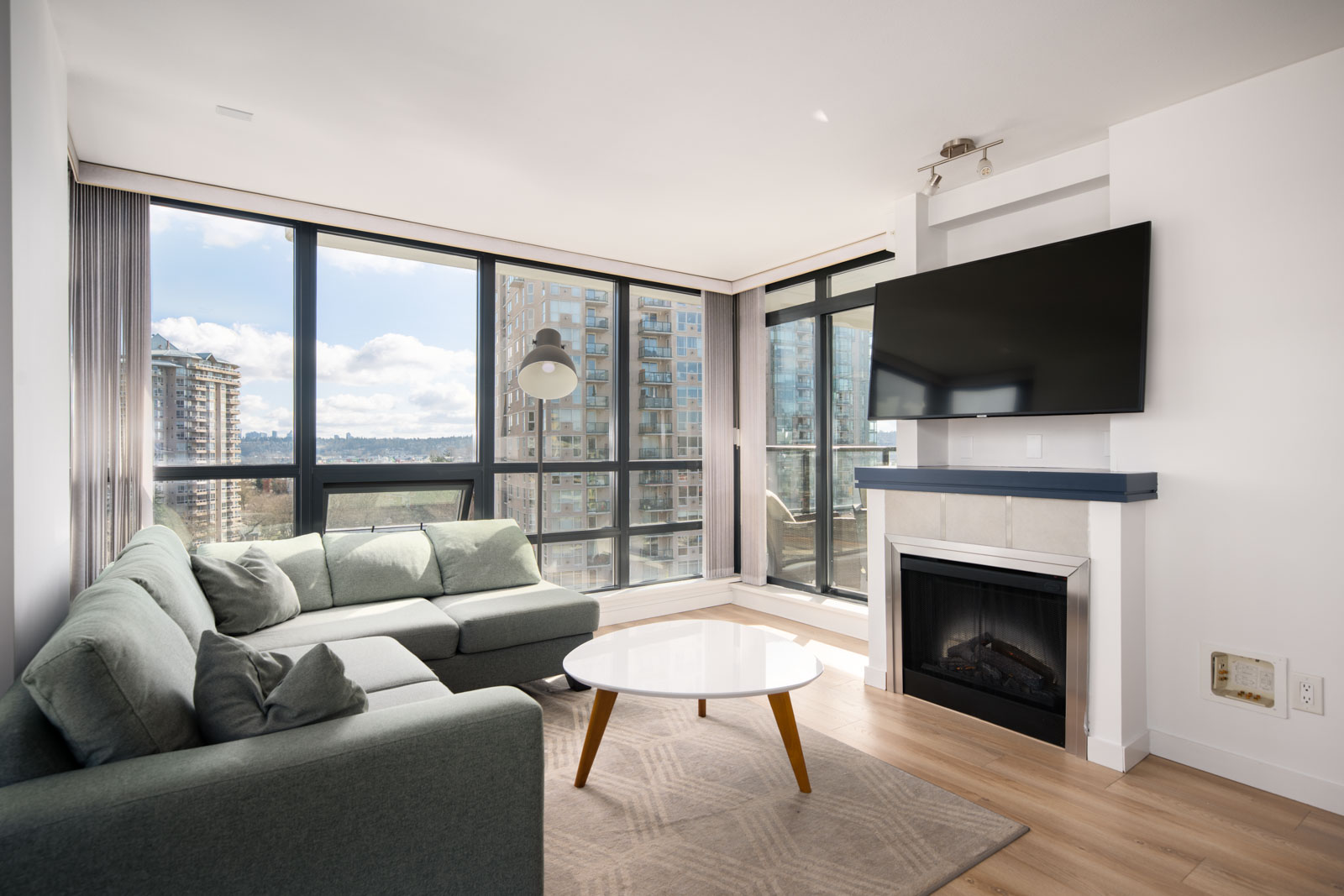 Modern living room with large windows, a grey sectional sofa, round white coffee table, wall-mounted TV, and fireplace, overlooking city buildings and a partly cloudy sky.