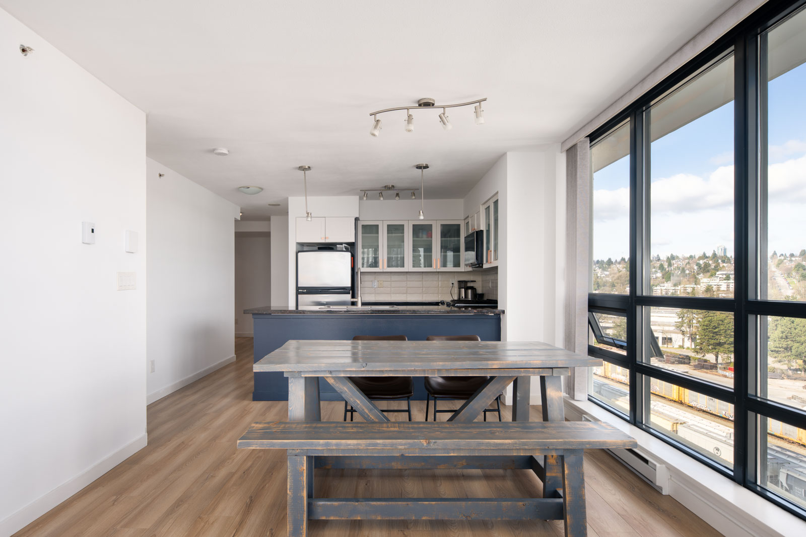A modern apartment dining area with a wooden table and bench, open kitchen with blue cabinets, large windows, and city views.