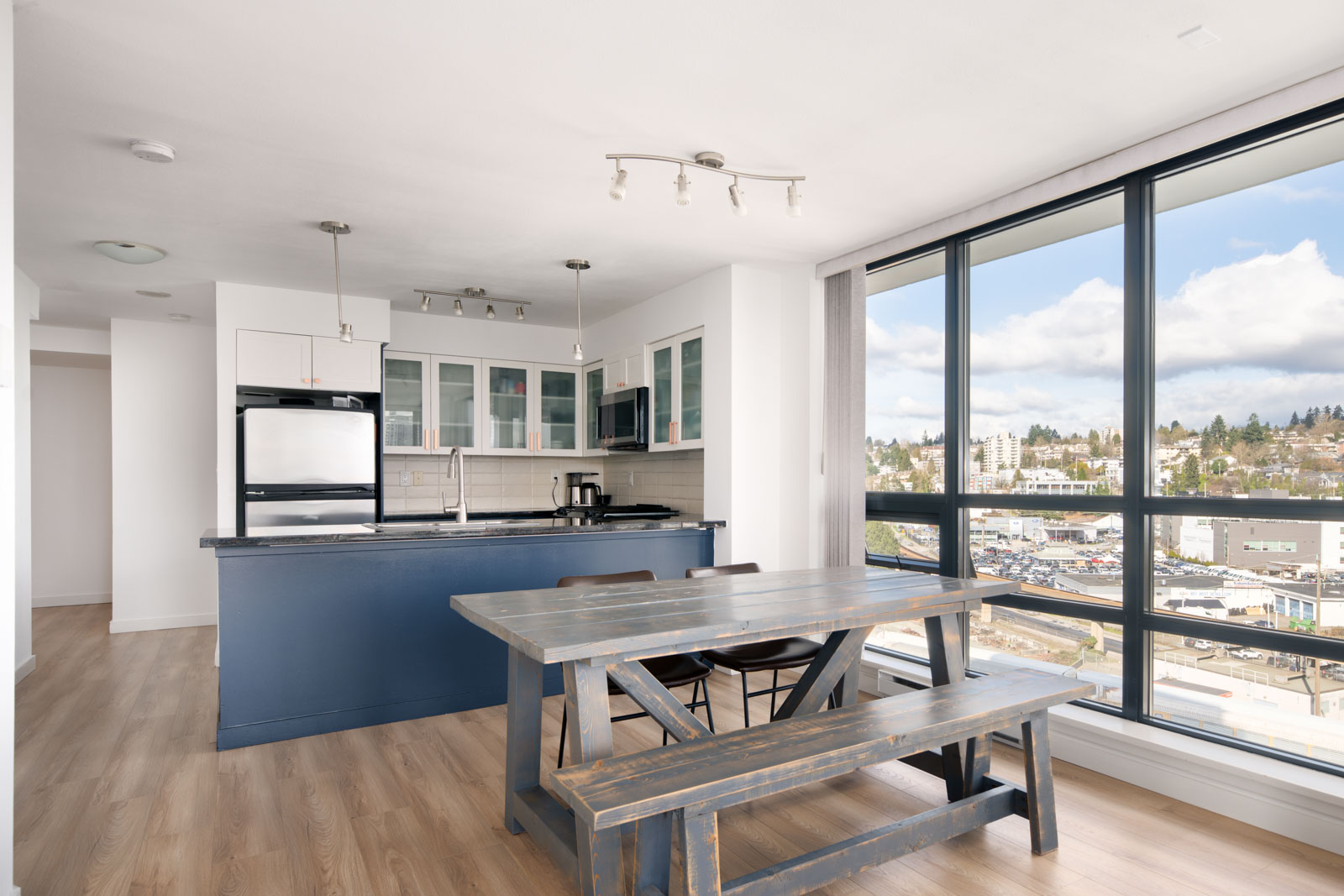 Modern kitchen and dining area with blue cabinetry, wooden table and benches, large floor-to-ceiling windows, and city view on a partly cloudy day.