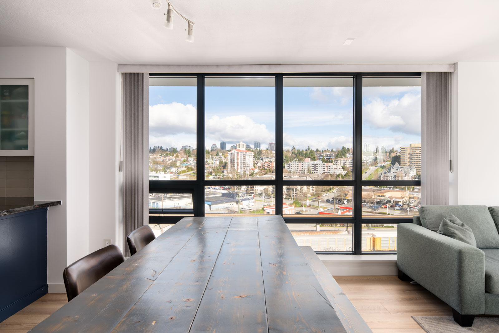 A modern dining area with a wooden table and chairs is set by large floor-to-ceiling windows overlooking a cityscape with buildings under a partly cloudy sky.