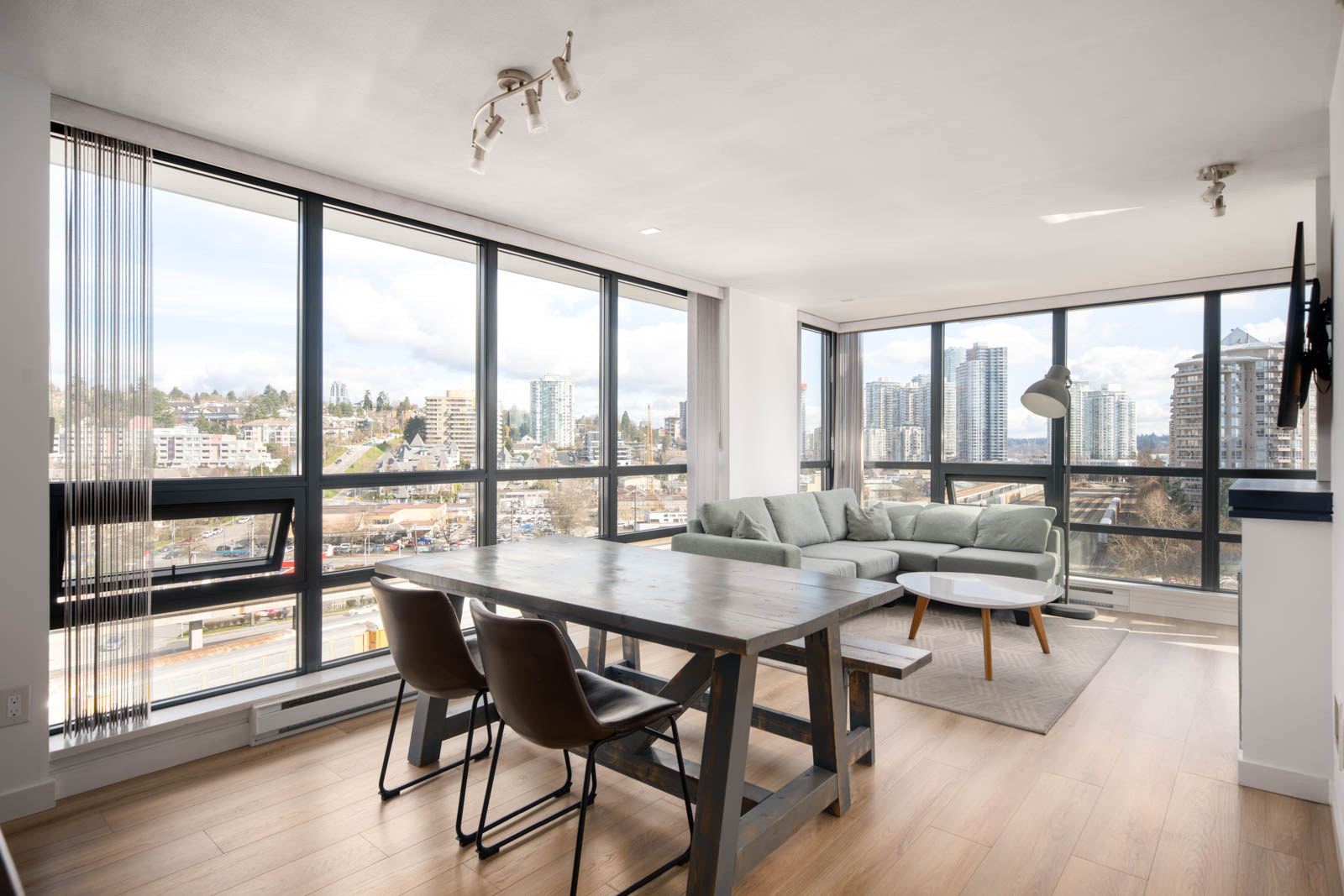 Modern apartment living room with large windows, a wooden dining table with chairs, a sofa, coffee table, and city skyline view in the background.