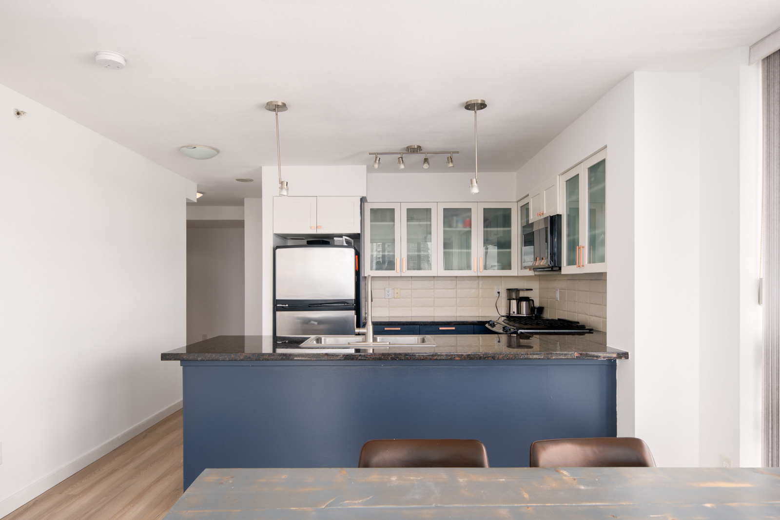 Modern kitchen with blue island, stainless steel appliances, glass-front cabinets, and a black countertop; dining table and chairs in the foreground.