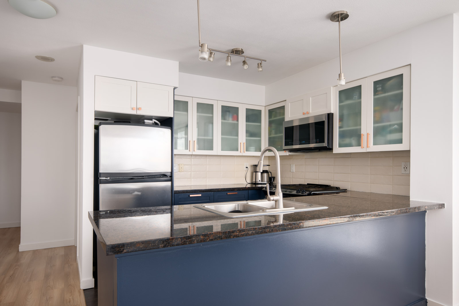 Modern kitchen with dark granite countertop, stainless steel appliances, white cabinets with glass doors, and a central sink island under pendant and track lighting.