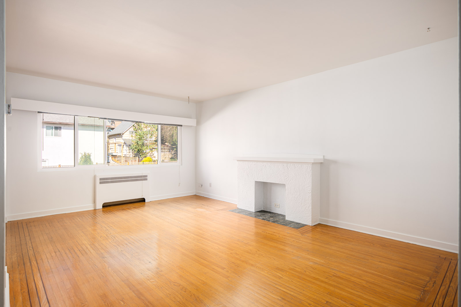 Empty living room with white walls, large window, hardwood floor, and a white fireplace.