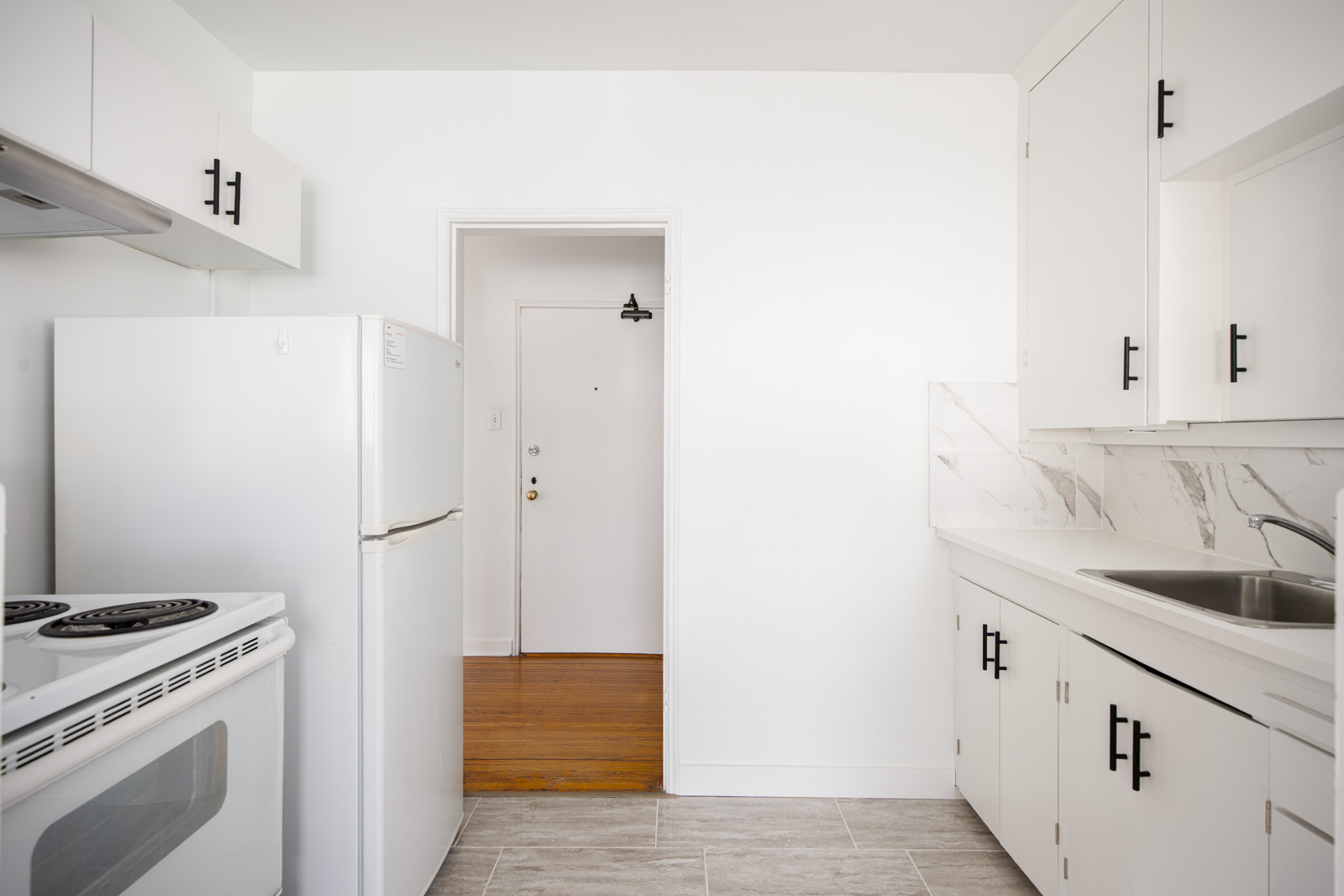 A small, white kitchen with an electric stove, refrigerator, sink, and white cabinets. The space features marble backsplash and light grey tile flooring. An open doorway leads to a different room.