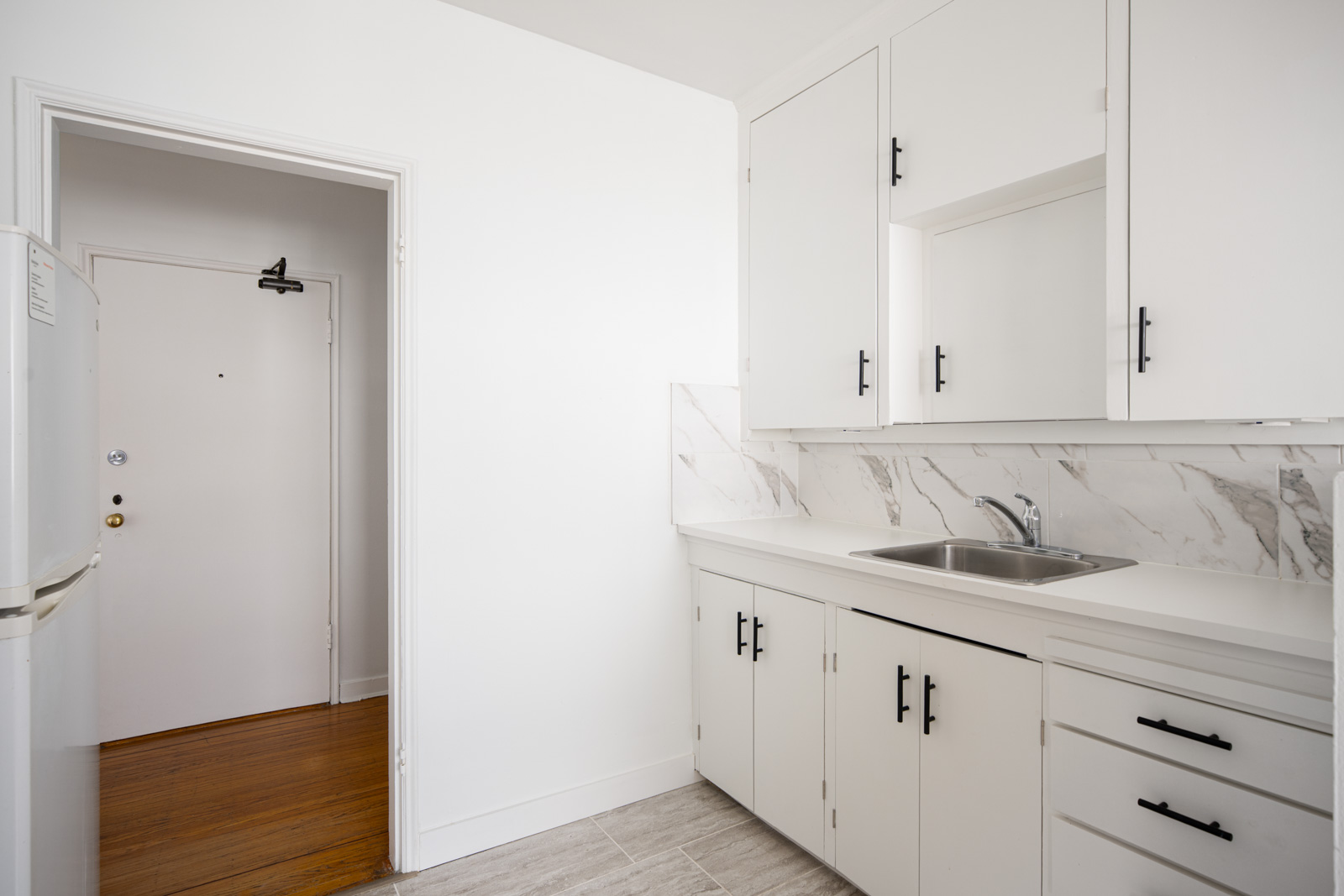 A small, clean kitchen with white cabinets, black handles, a stainless steel sink, marble backsplash, and a view of an open doorway leading to a hallway.