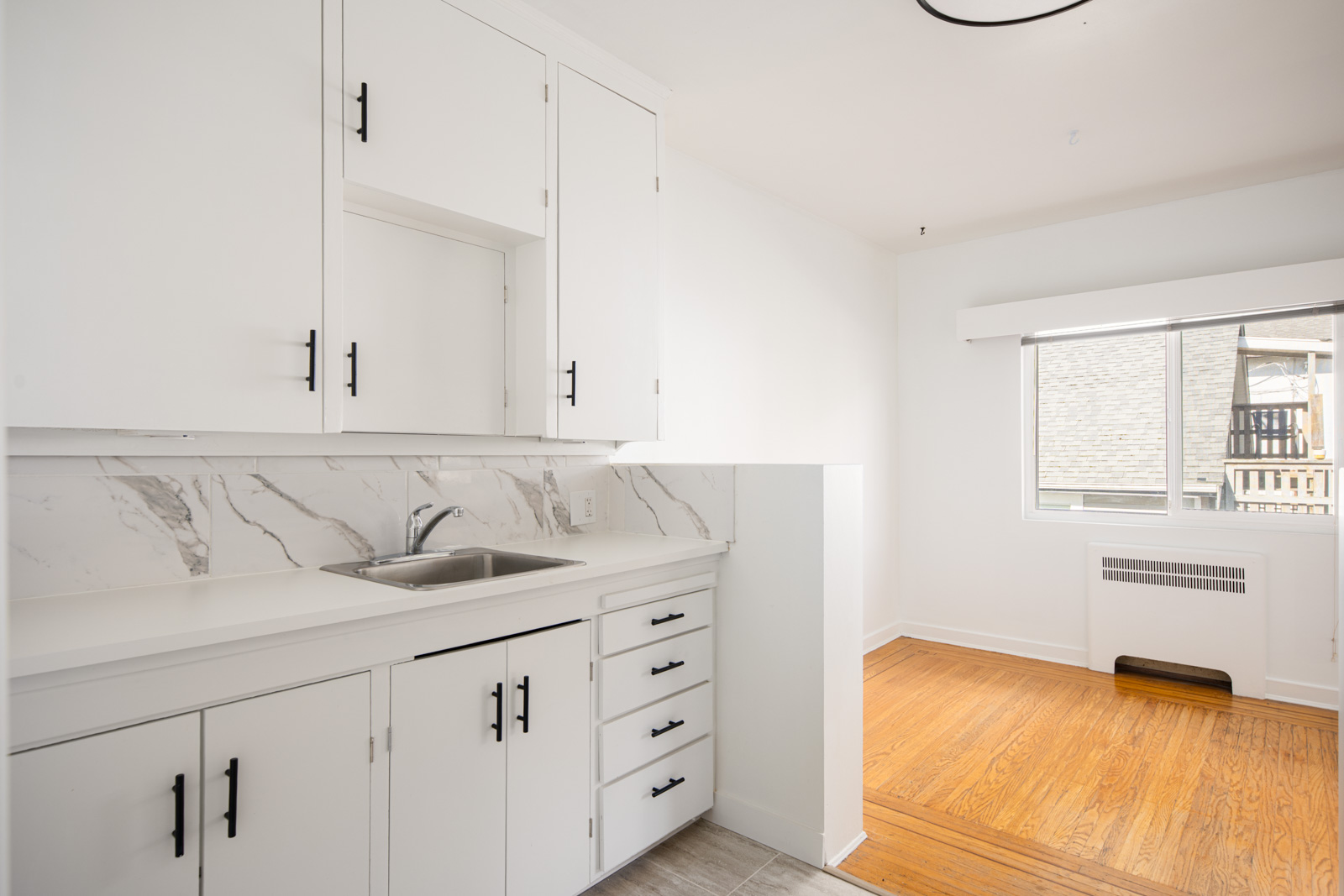 A small kitchen with white cabinets, marble backsplash, stainless steel sink, and an adjacent area with wood flooring and a large window.